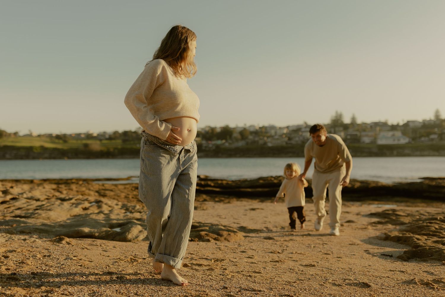 A pregnant woman stands on a sandy beach while a man holds a toddler's hand near a calm waterway at sunset.