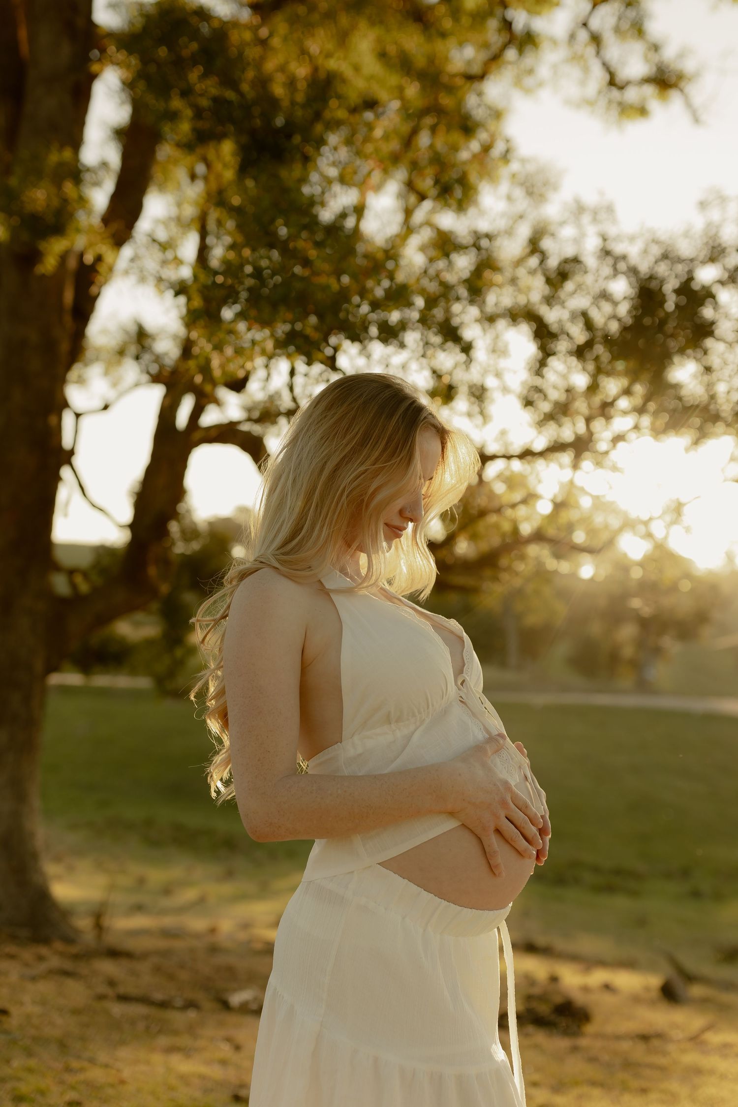 Pregnant woman in white dress cradling baby bump outdoors in golden sunset light near large tree.