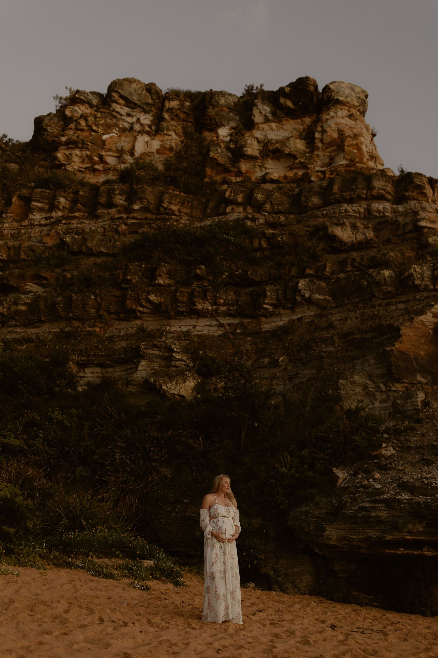 Pregnant woman in white lace dress stands on sandy ground before dramatic rocky cliff face surrounded by lush greenery.