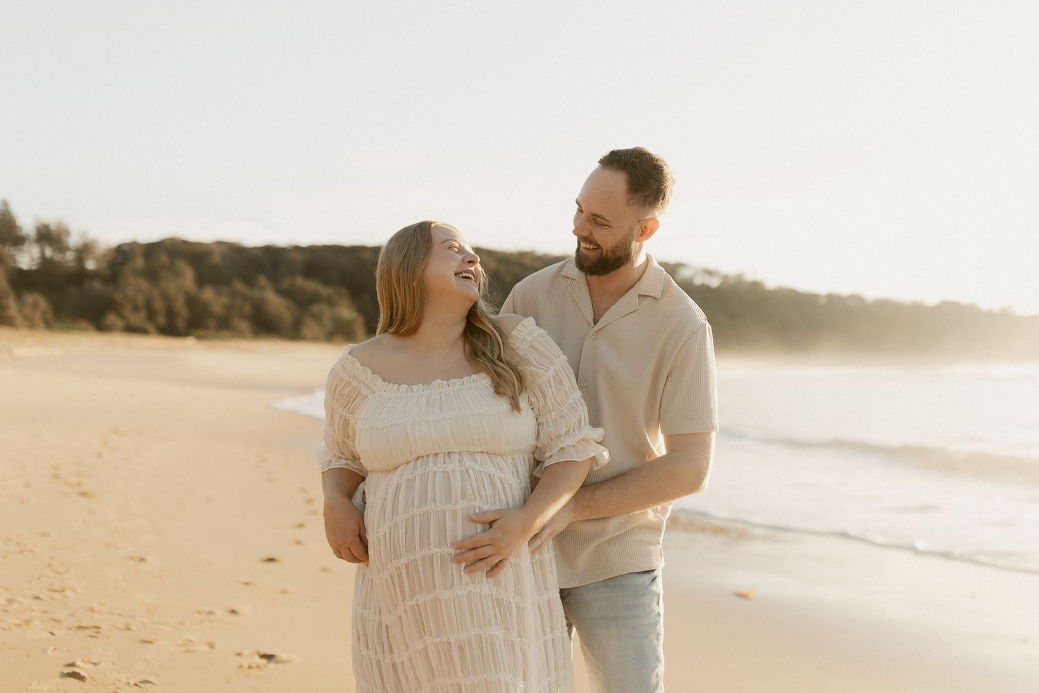 Pregnant couple smiling and walking on a sunny beach, woman in white lace dress cradling baby bump.
