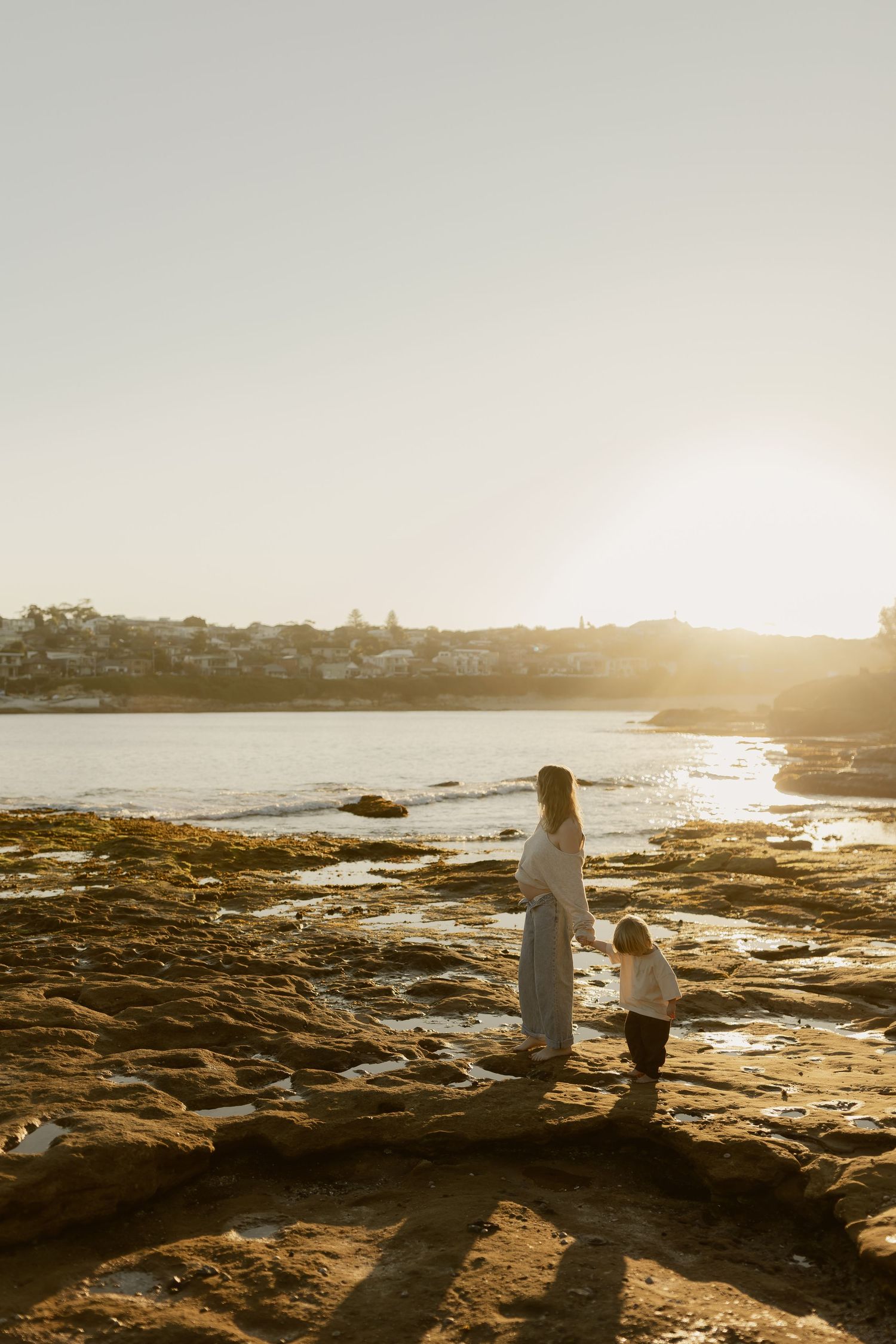 Mother and child standing on rocky shore at golden sunset with coastal town in background.