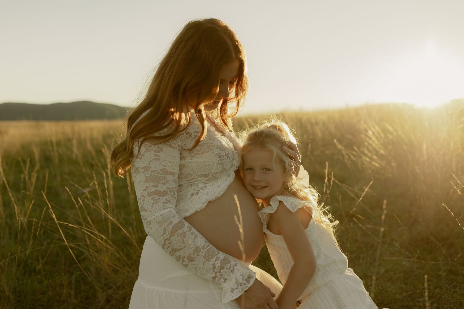 A mother and young daughter in white lace dresses embrace in a golden sunlit field at sunset.