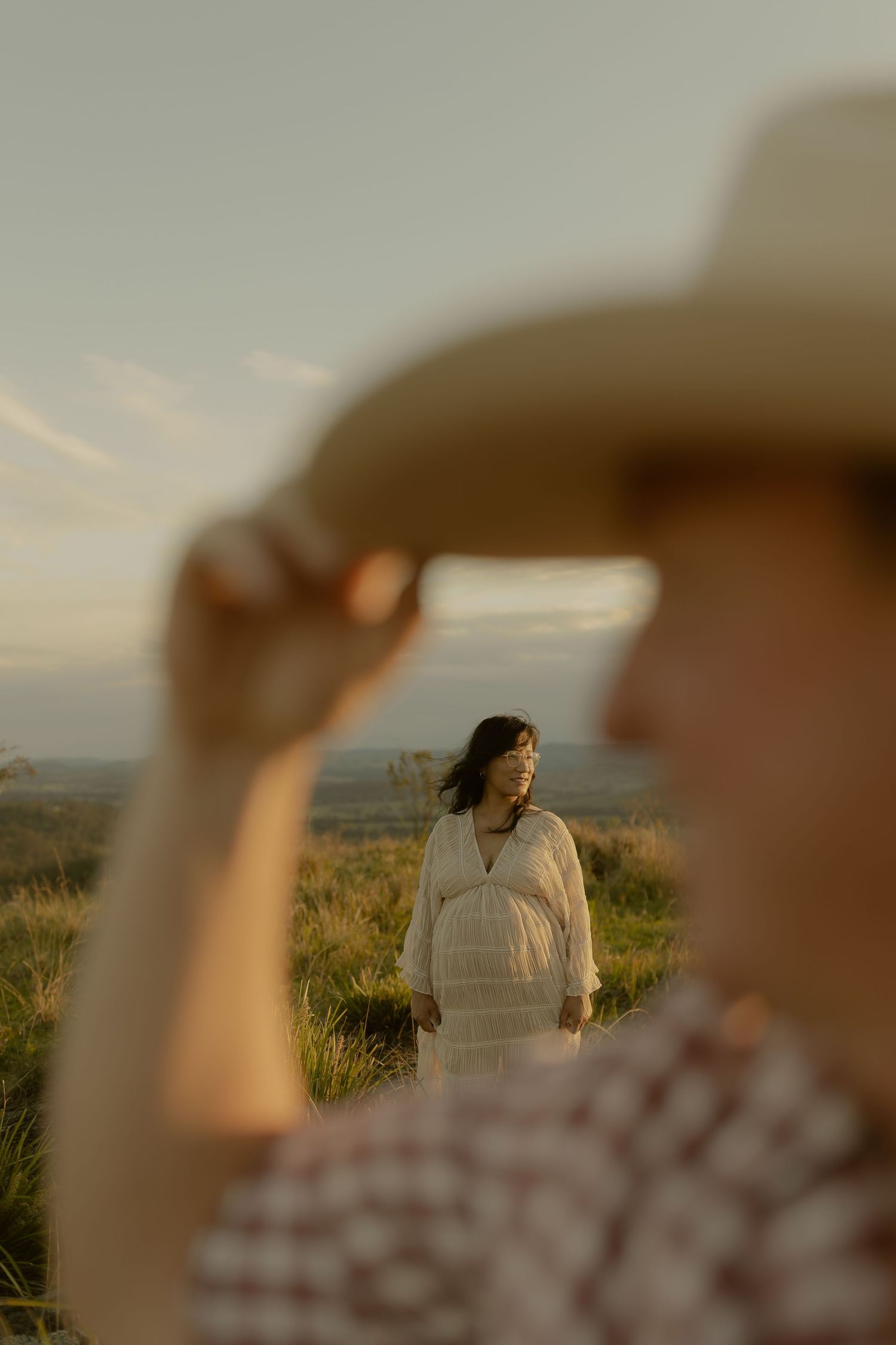 A woman in a white dress stands in golden fields at sunset, framed by a man tipping his cowboy hat in the foreground.