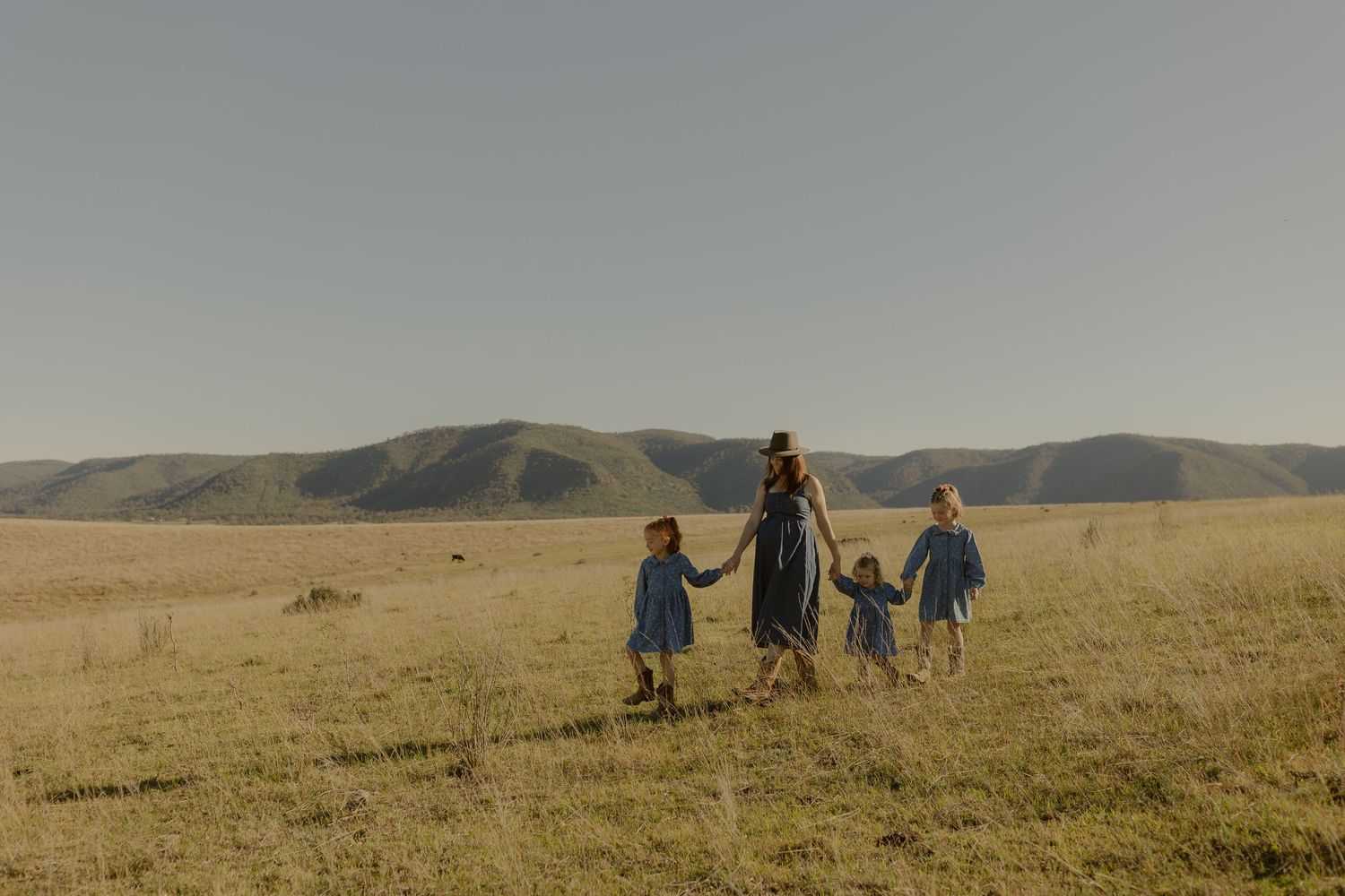 Mother walking hand in hand with three children in blue dresses across a sunny open field with hills behind.