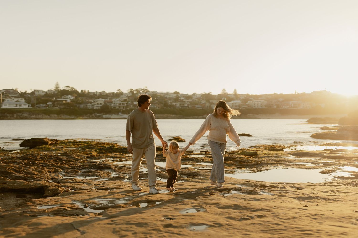 Family of three holding hands walking on rocky beach at golden sunset with coastal town in background.
