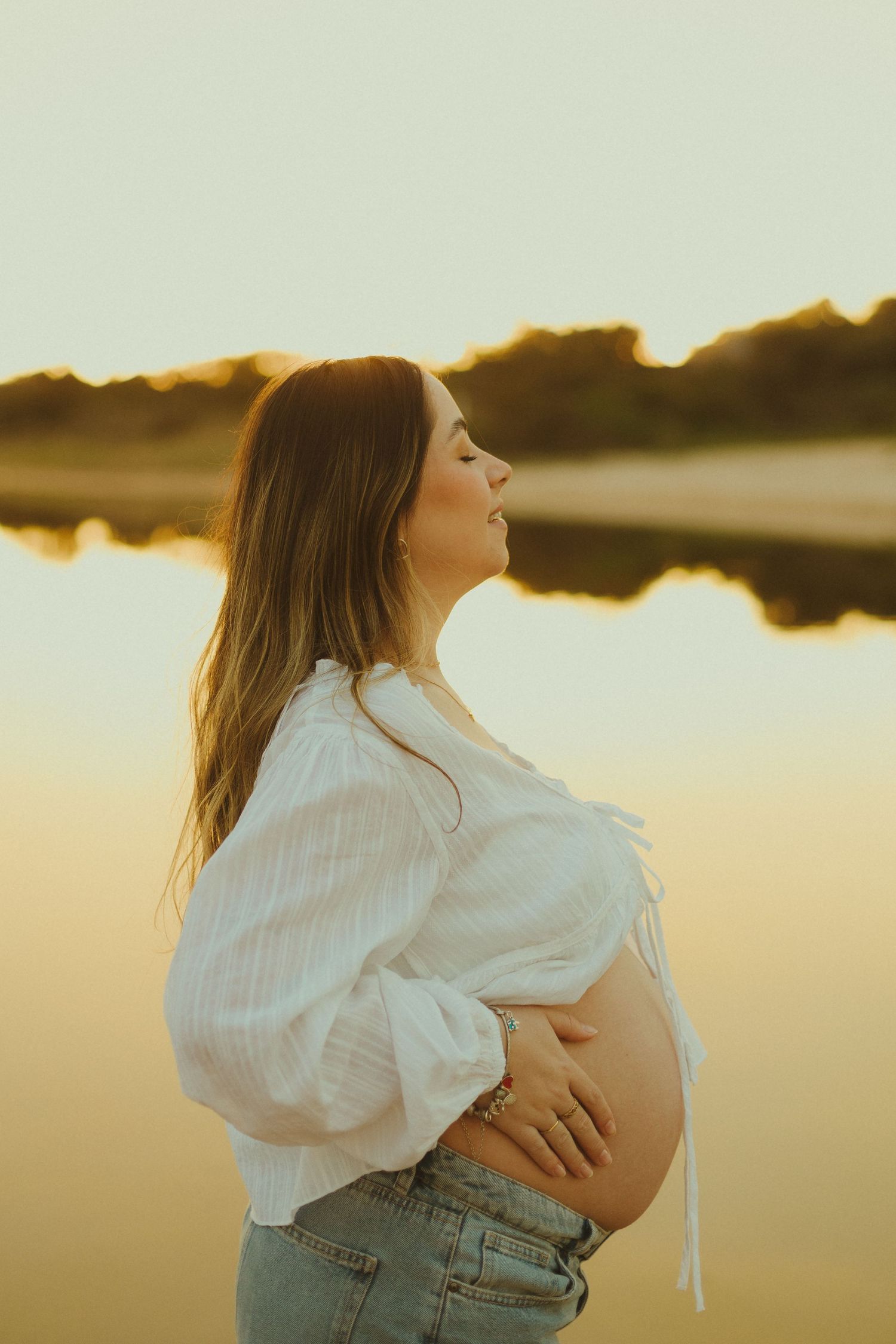 Expectant mother in a white blouse caresses her pregnant belly during golden sunset by tranquil lake.