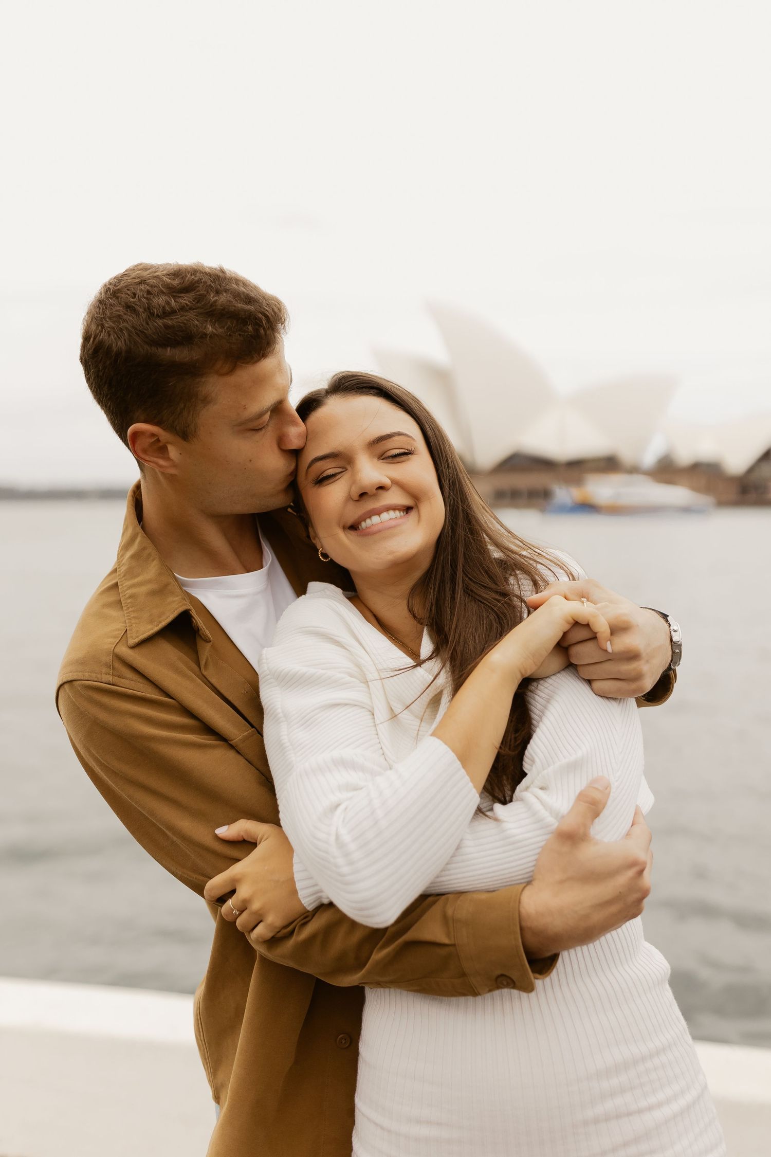 A romantic couple embraces near Sydney Opera House with one person in brown coat and other in white sweater.