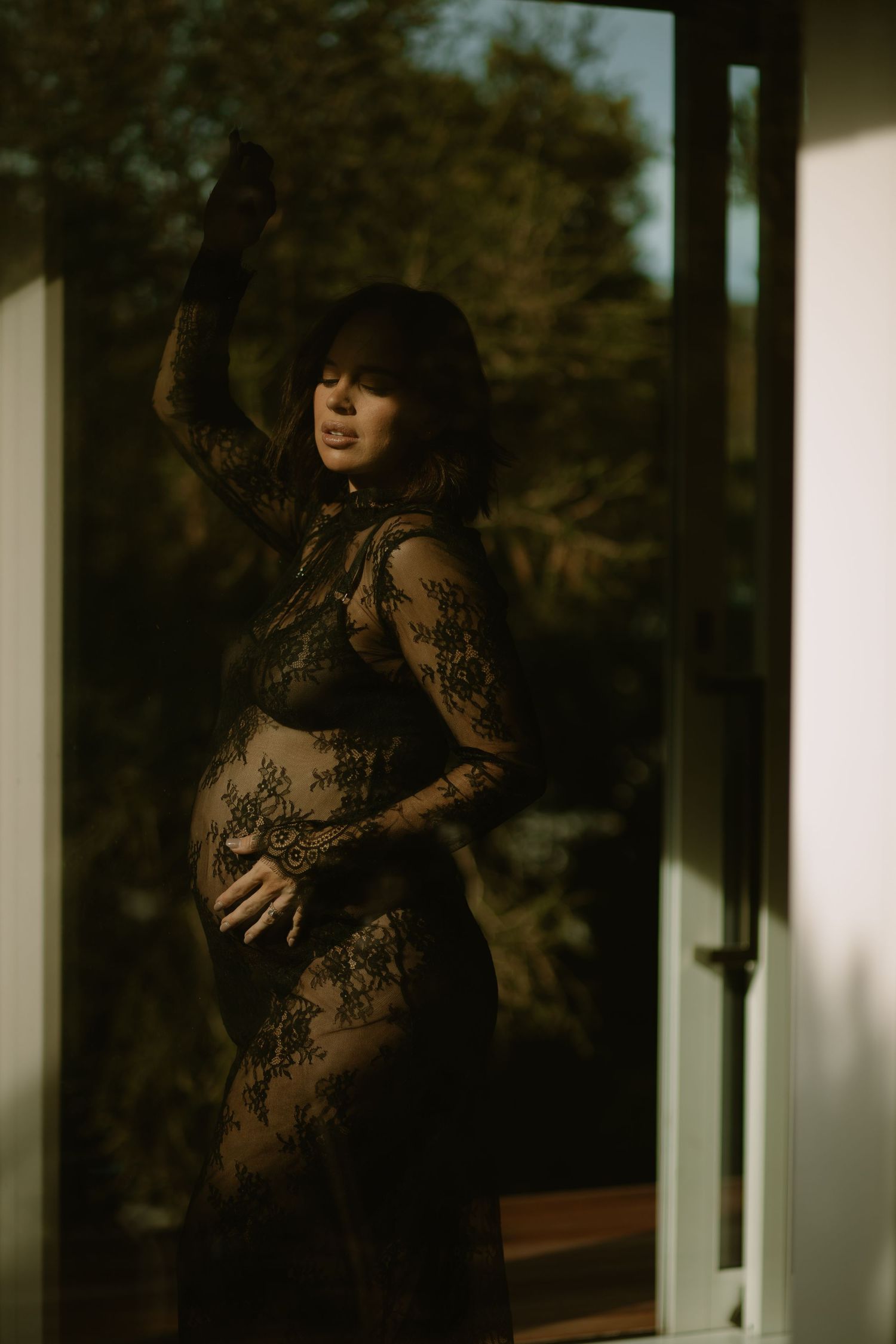A maternity photoshoot featuring a black lace dress in warm natural lighting by a window.