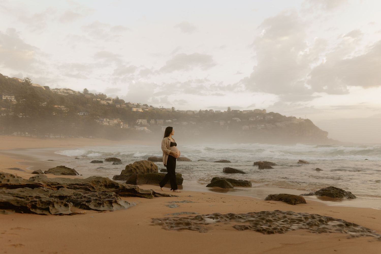 A solitary figure stands on rocky coastal beach during misty sunset with waves crashing on the shore.