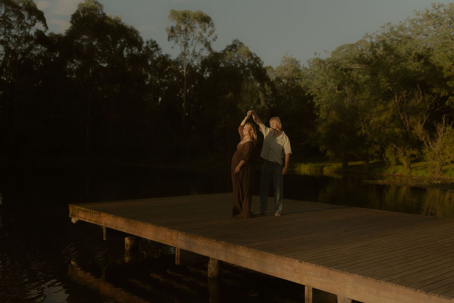 Long wooden pier extends over still waters with trees lining the shore at dusk.