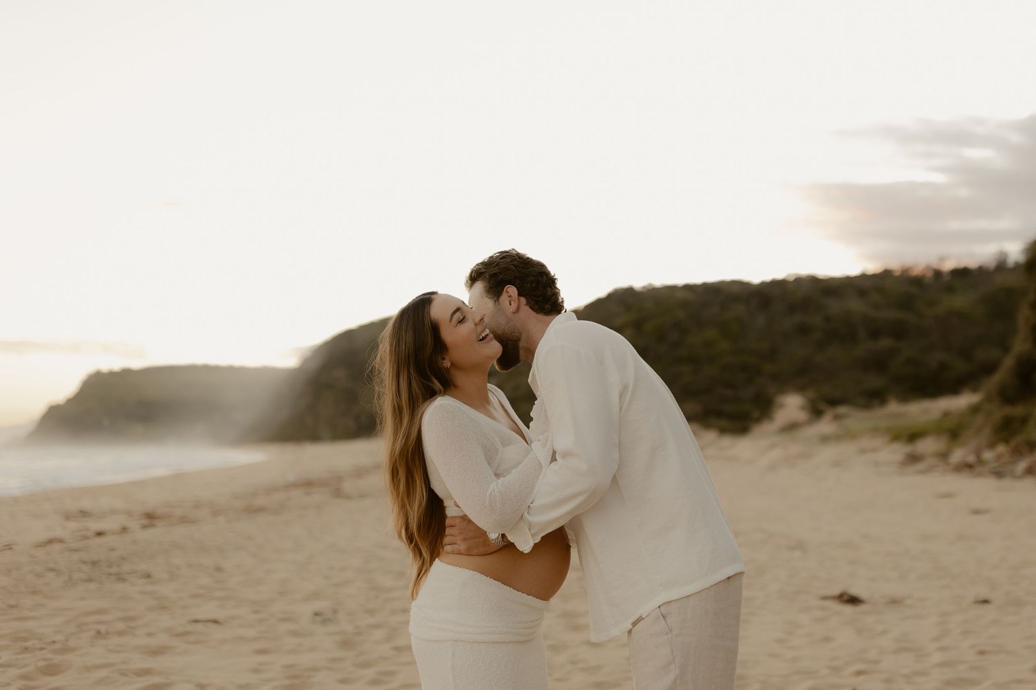 A couple shares a romantic kiss on a sandy beach during sunset with waves crashing in the background.