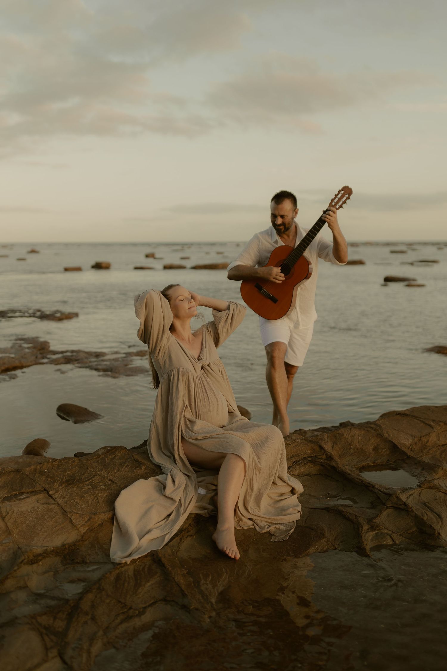 Pregnant woman in flowing beige dress sits on coastal rocks while man plays acoustic guitar at sunset.