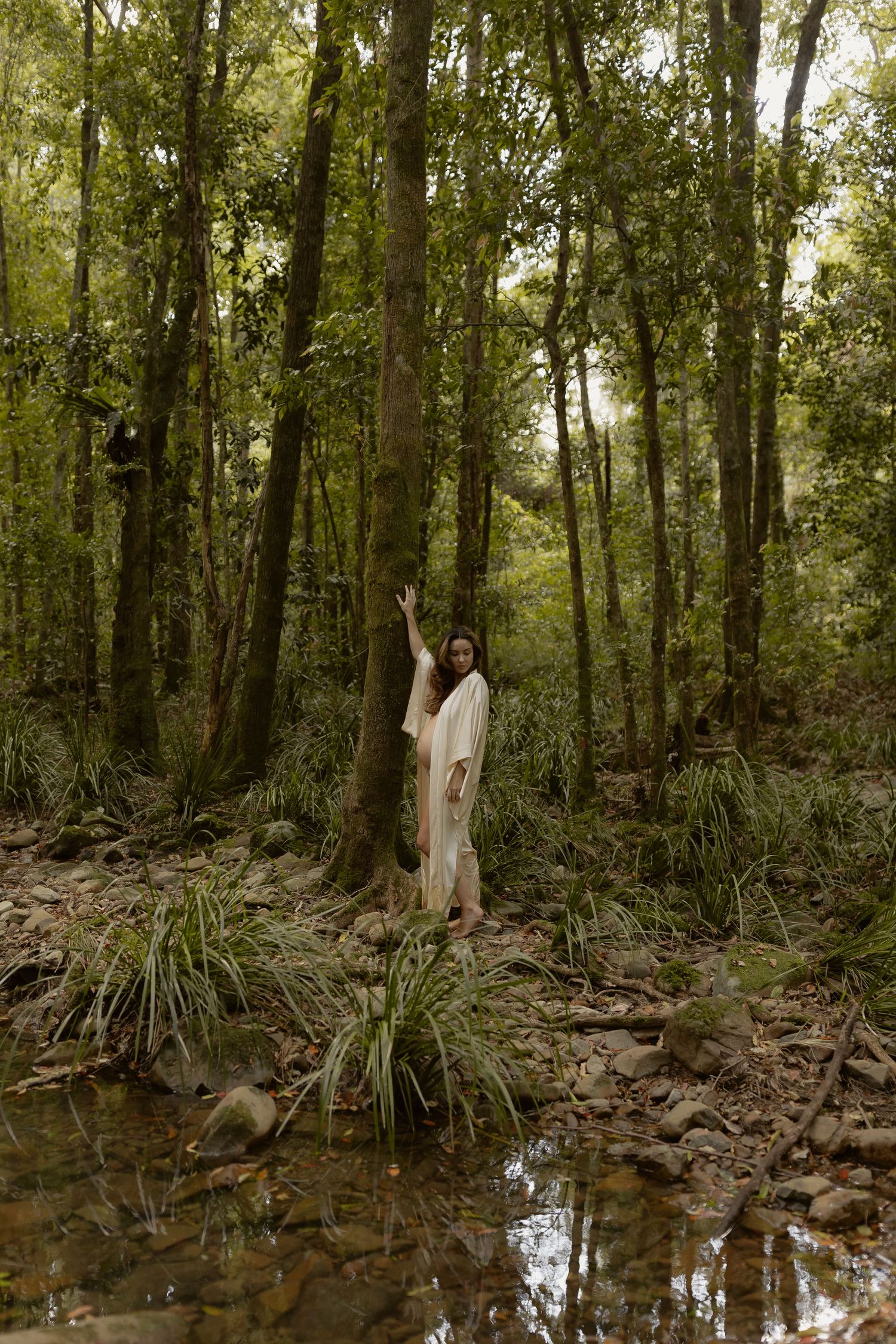 Woman in white robe standing by a rocky stream in a lush green forest, reaching up to touch a tall tree.