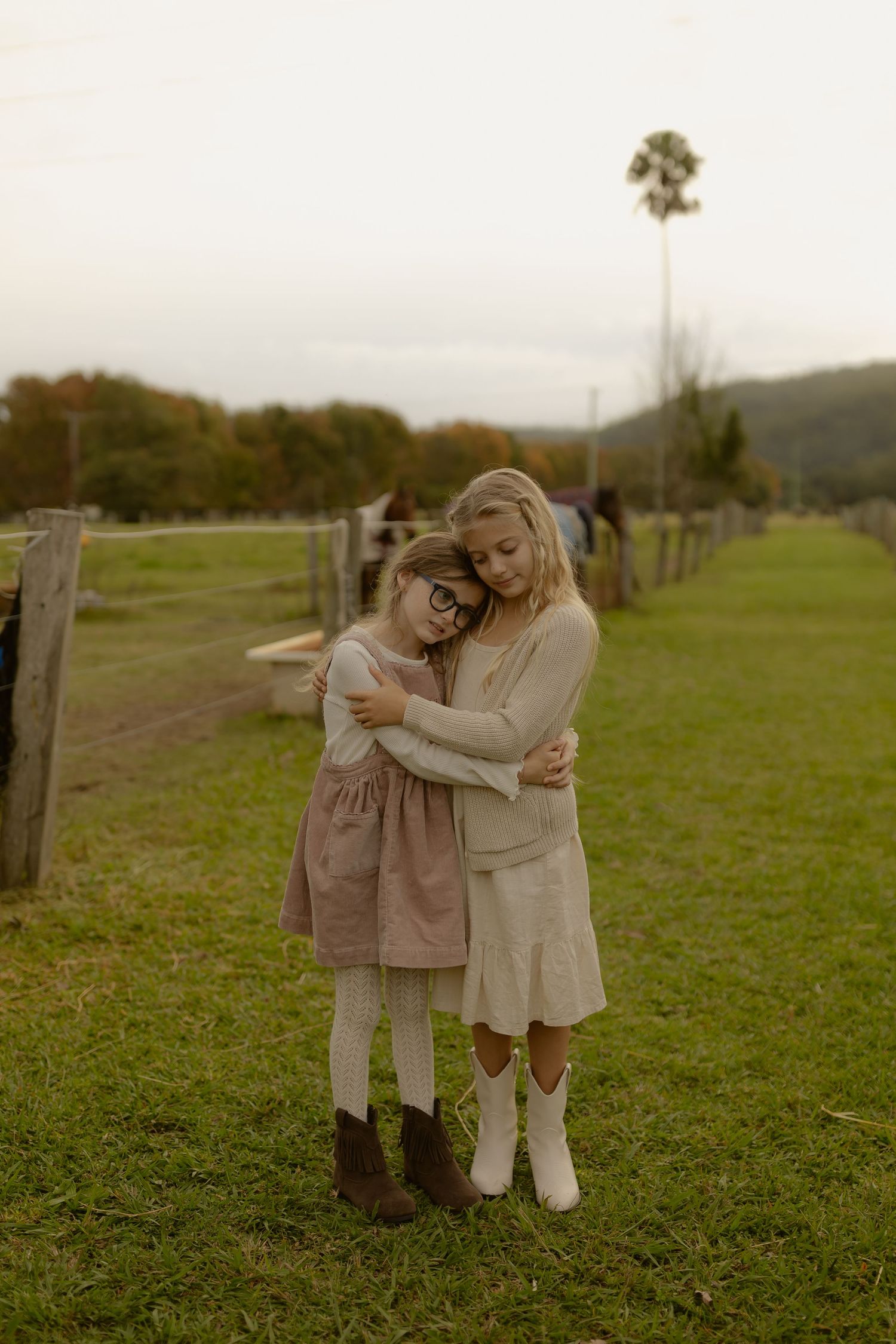 Two young girls in vintage-style dresses share a tender hug on a green farm paddock.