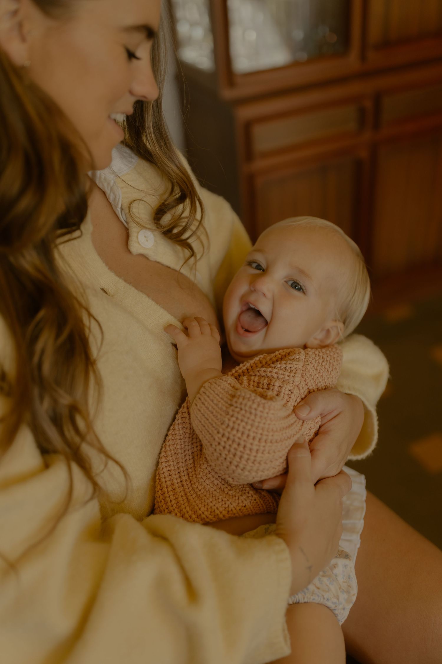 A smiling mother in a yellow cardigan holds her laughing baby dressed in a brown knit romper indoors.