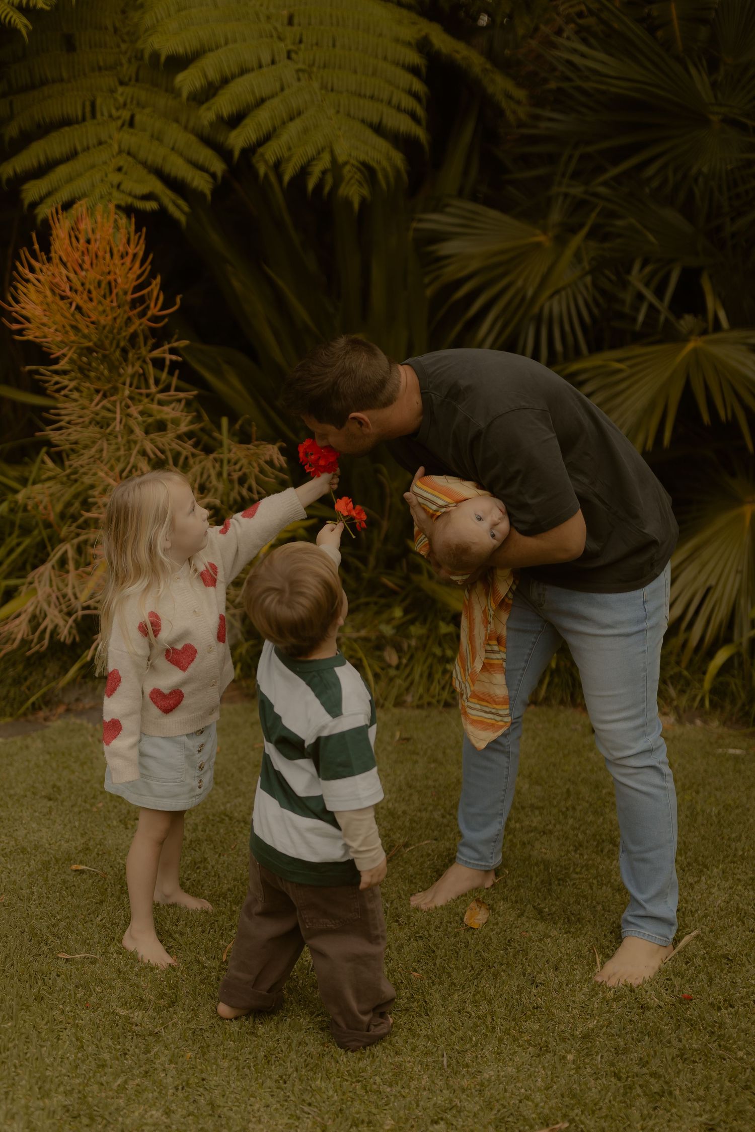 Father bending down to smell red flowers offered by blonde girl while two young children stand in tropical garden.
