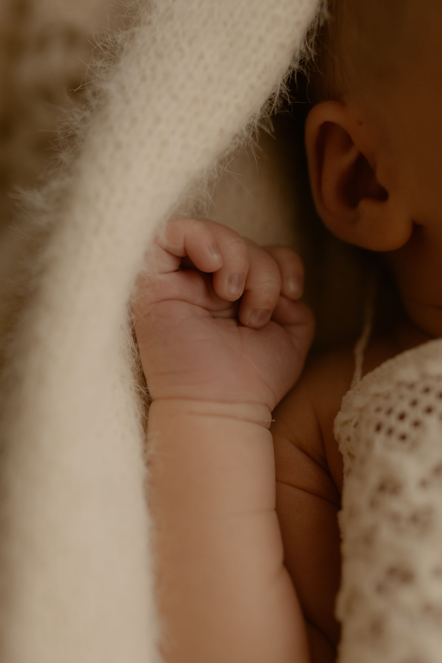 Newborn baby's tiny hand grasping against a soft white blanket in warm lighting.