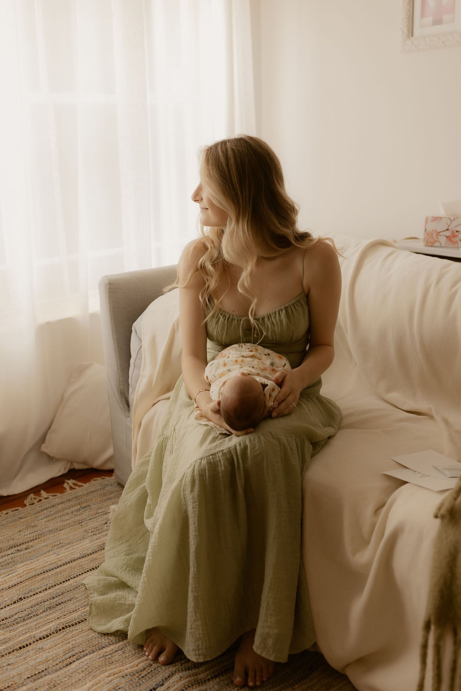 Mother in sage green dress sitting on white couch holding newborn baby in natural light.