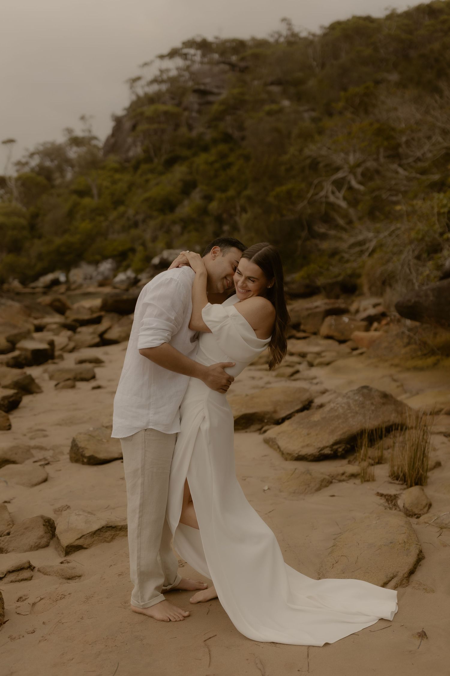 A couple in white attire shares romantic moments on rocky coastal terrain at sunset.