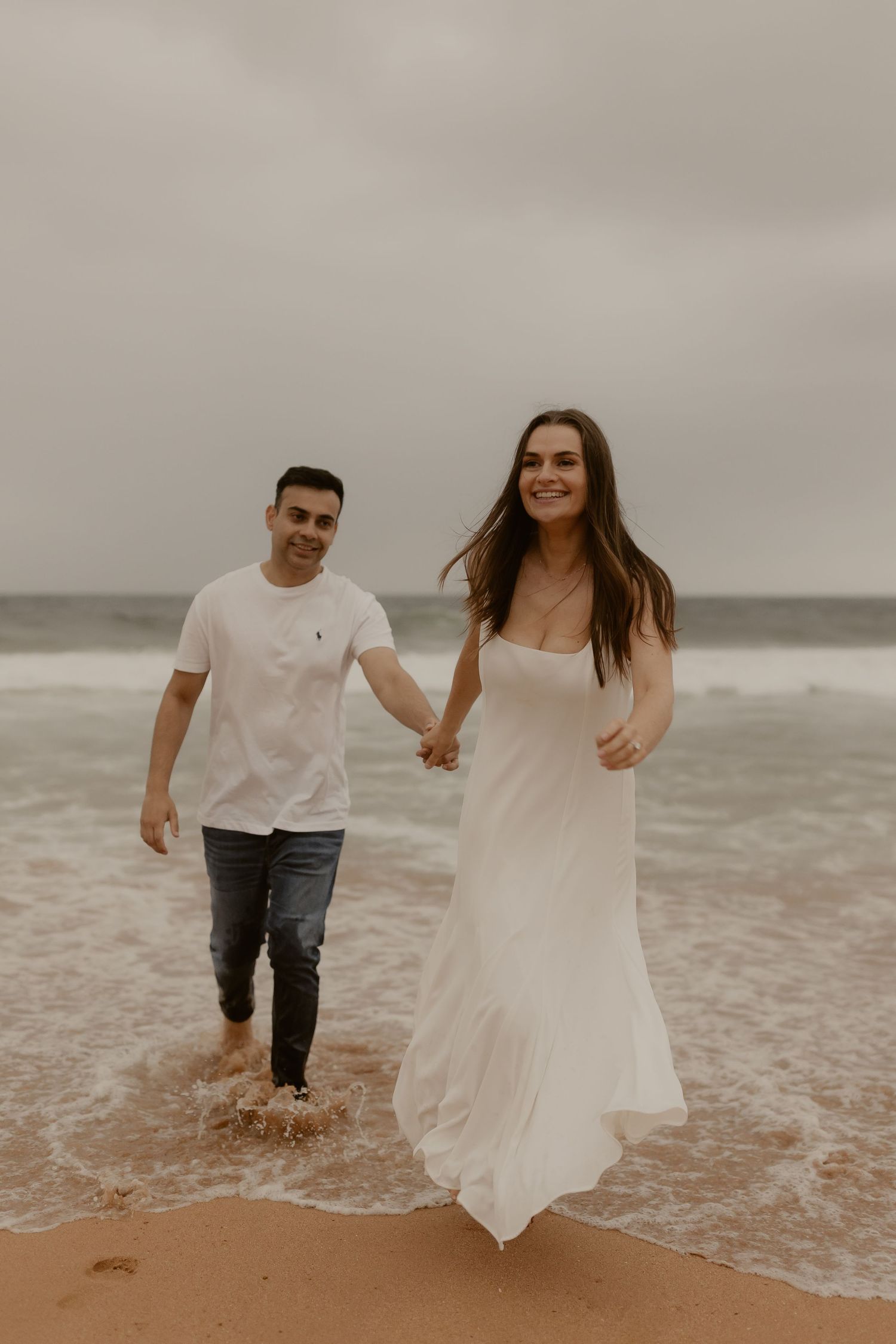 A couple plays and splashes in the ocean waves, with the woman's white dress flowing in the wind.
