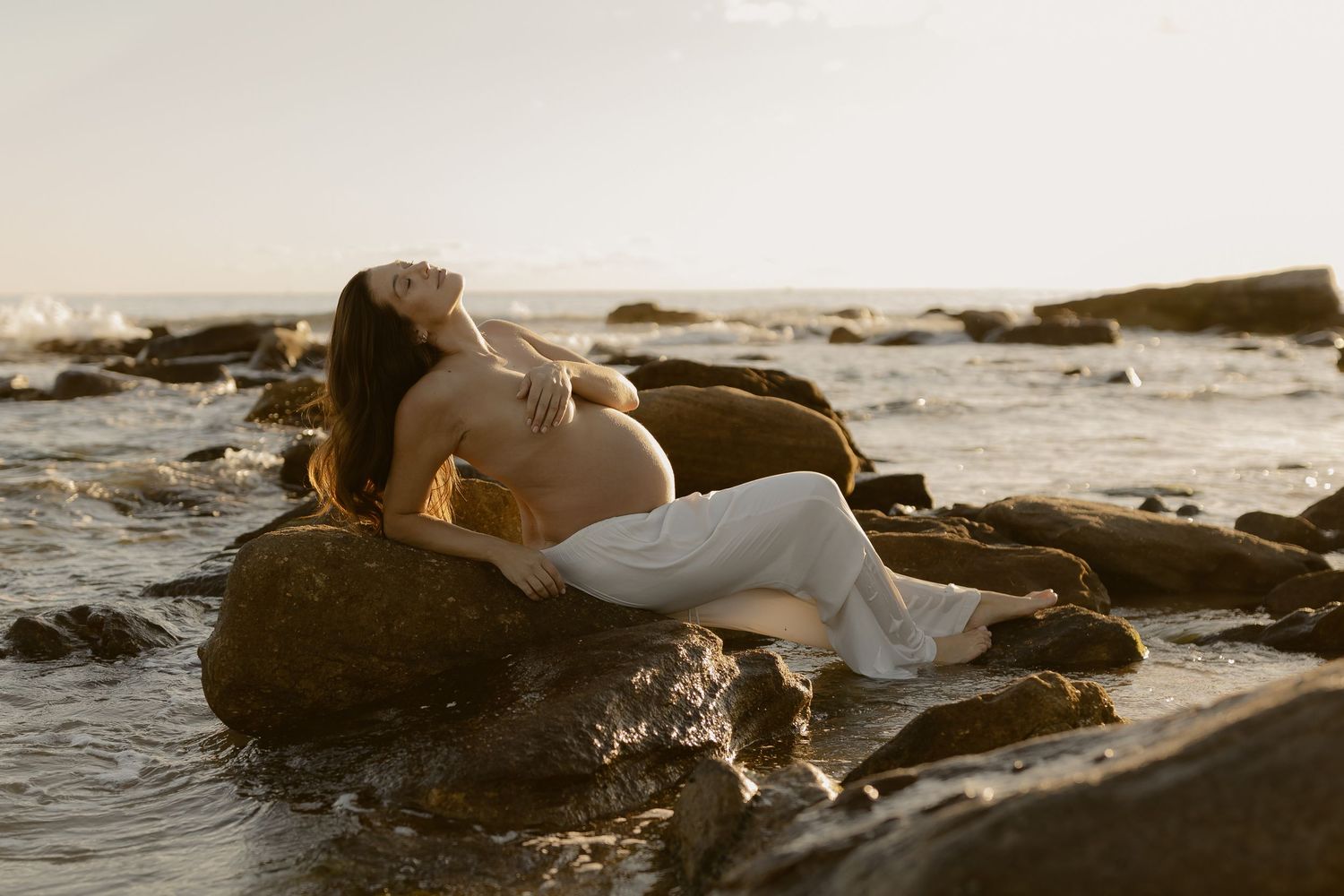 A couple embraces while lying on rocks near the ocean during sunset creating a romantic silhouette.