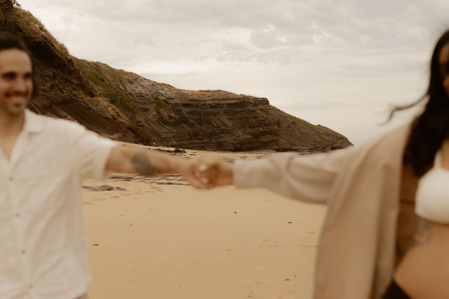 A couple embraces on a scenic beach with rocky cliffs and cloudy skies in the background.
