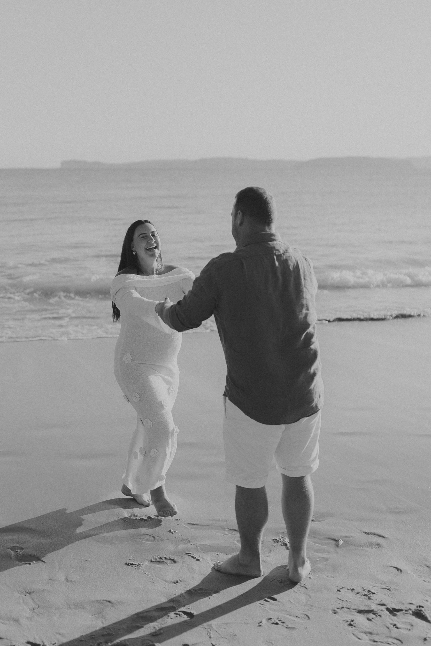 A couple shares a playful moment together on a misty beach, captured in black and white.