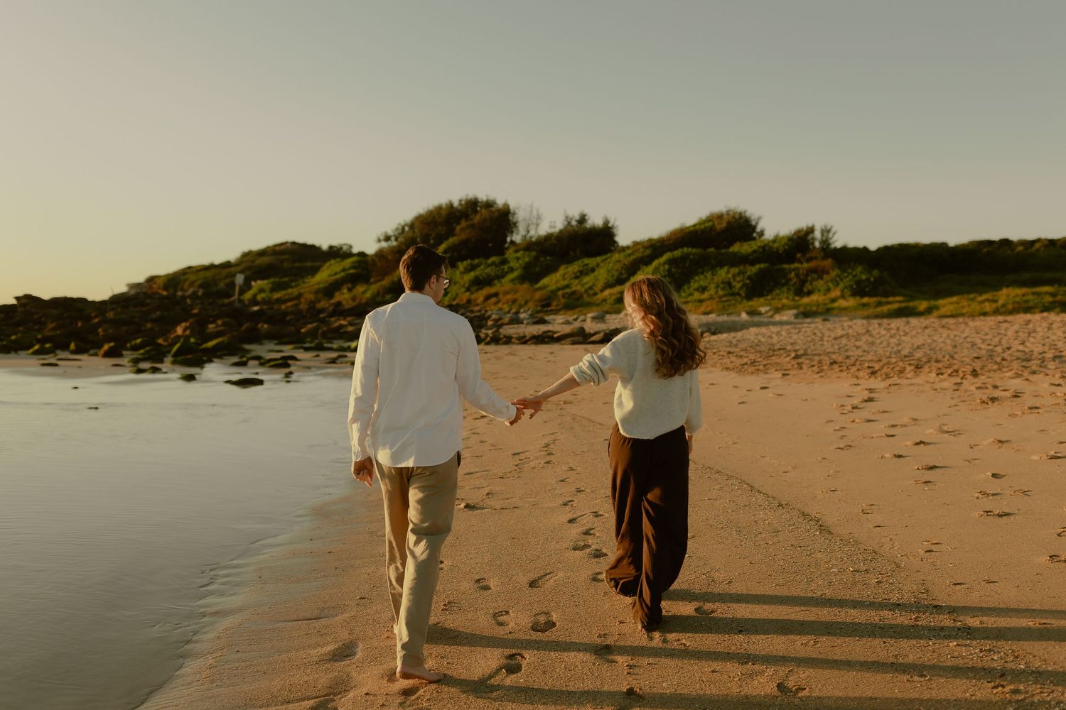 Couple holding hands walking along sandy beach at sunset, wearing white shirts and dark pants.