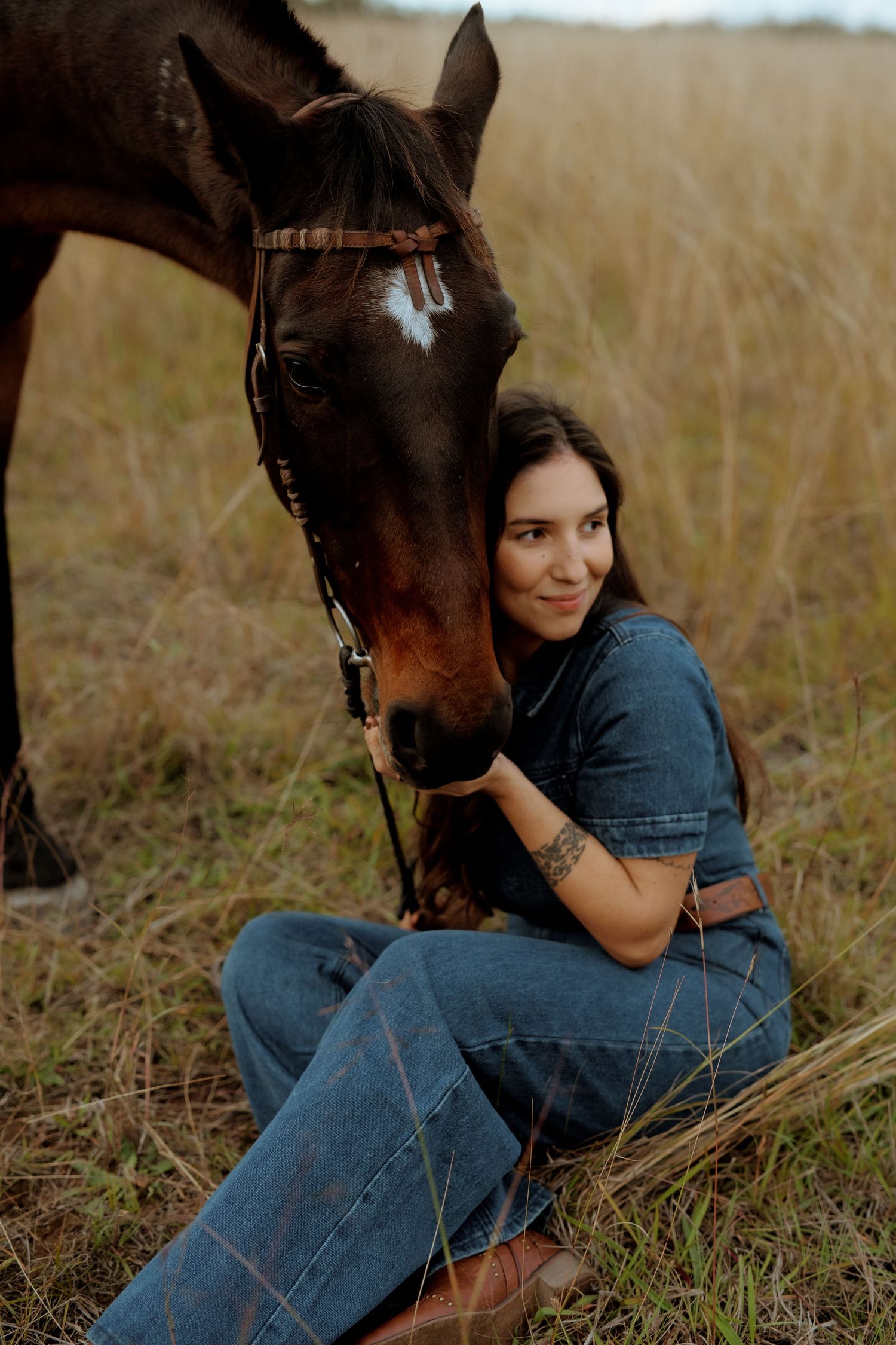 Woman in denim sitting in a field, affectionately leaning against a brown horse.
