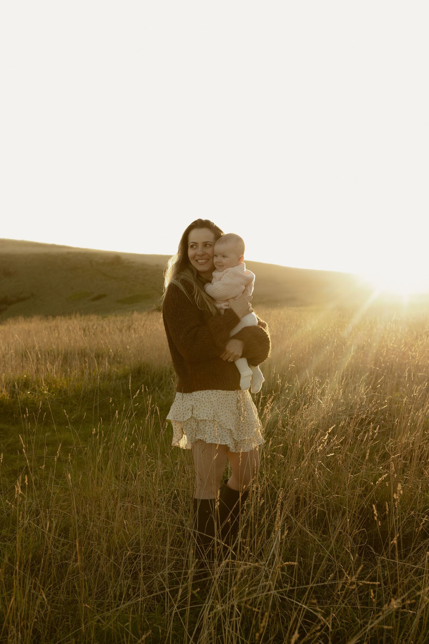 A smiling mother holds her baby in a sunlit golden field at sunset, wearing a dark sweater and floral skirt.