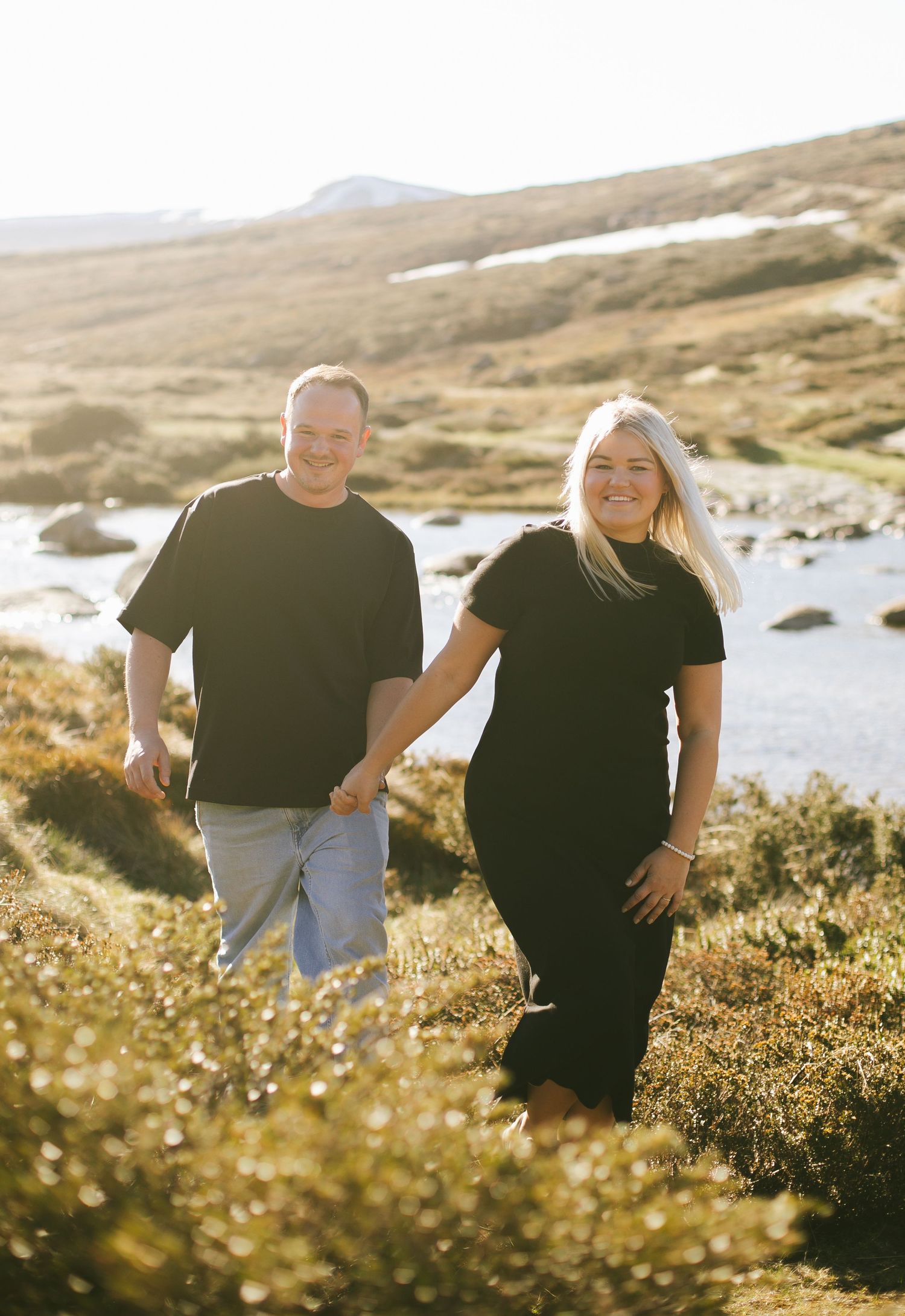 A couple in matching black t-shirts hold hands while standing in a sunlit mountainous landscape by the water.