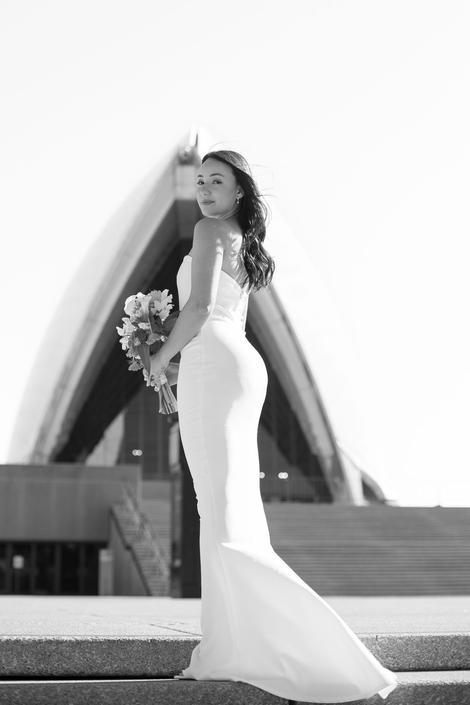 Elegant bride in fitted white mermaid wedding dress poses on steps with bouquet in black and white photography.