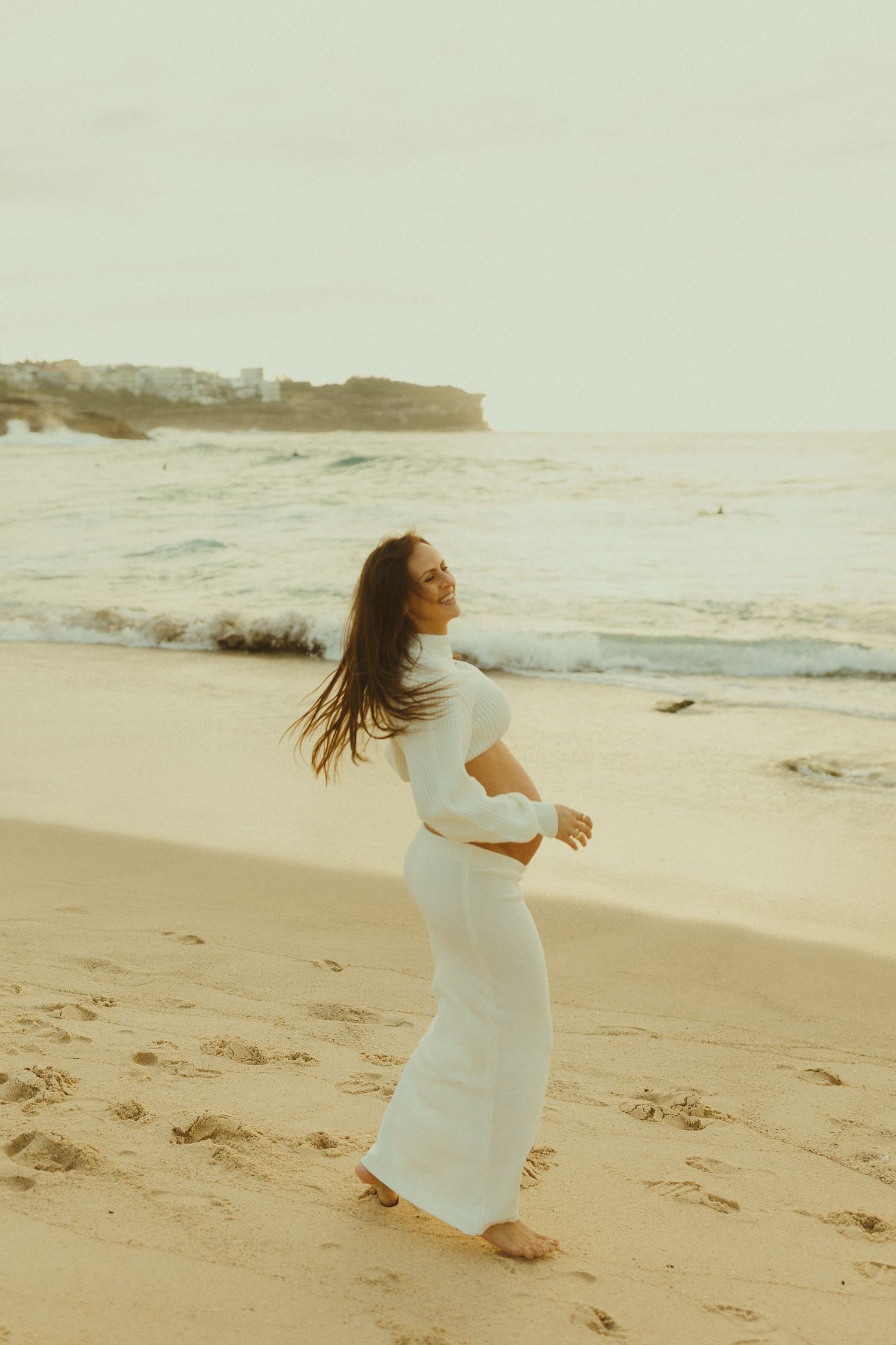 A person in a flowing white dress dances joyfully on a sandy beach at sunset.