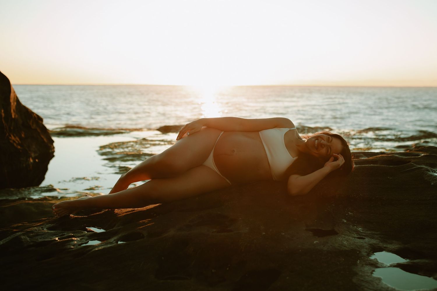 Person in white swimsuit lying on rocky beach at sunset with sunlit ocean horizon in background.