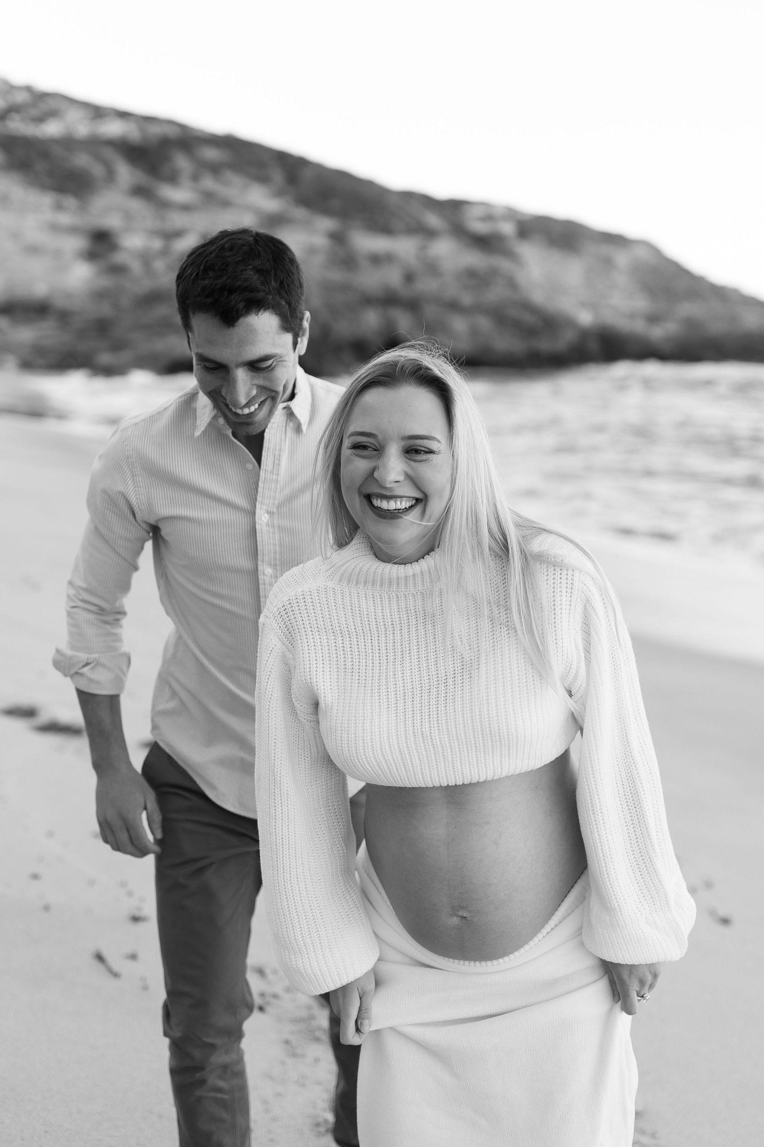 Black and white photo of a couple at the beach, with her wearing a white sweater showing off her pregnant belly.