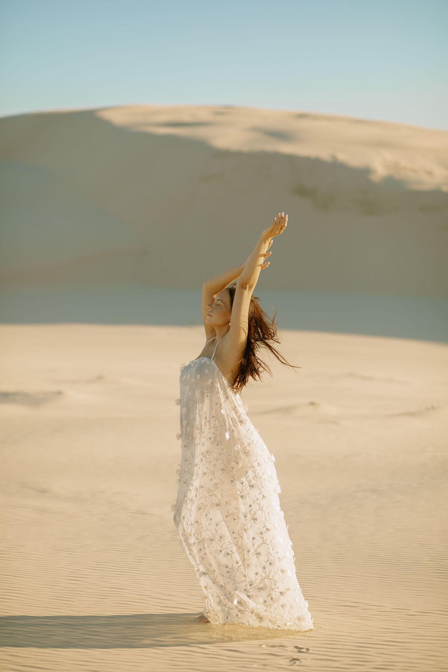 Woman in flowing white lace dress spins gracefully across desert sand dunes during a golden sunset.