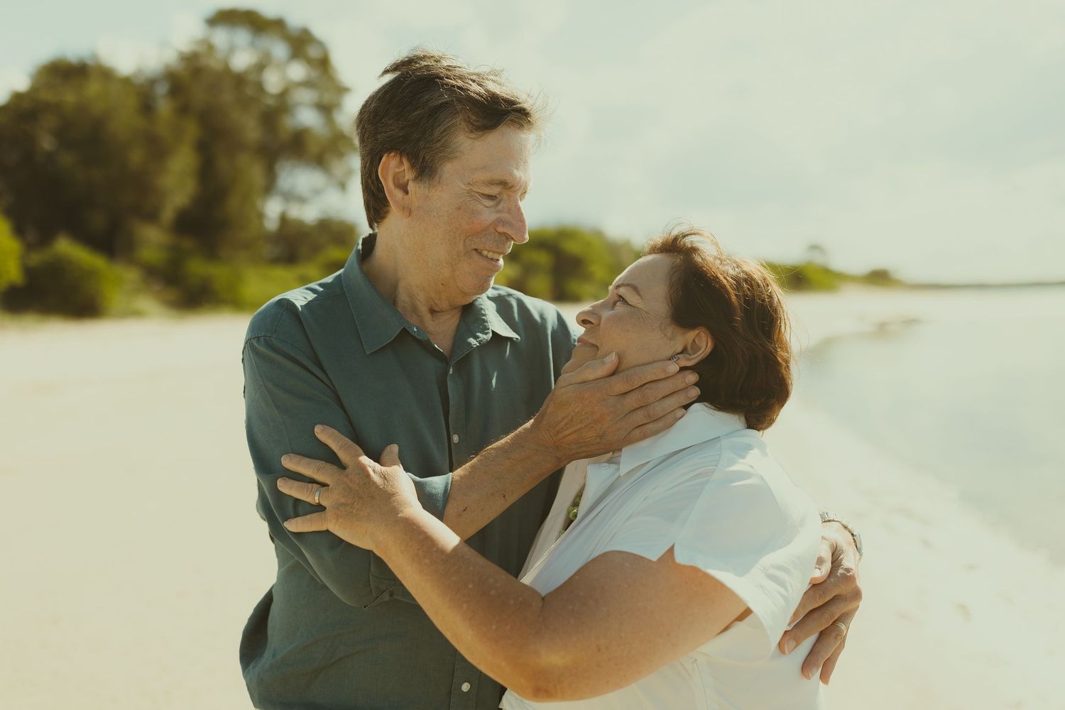 A couple shares a tender moment while walking along a sunny beach with white sand and palm trees.