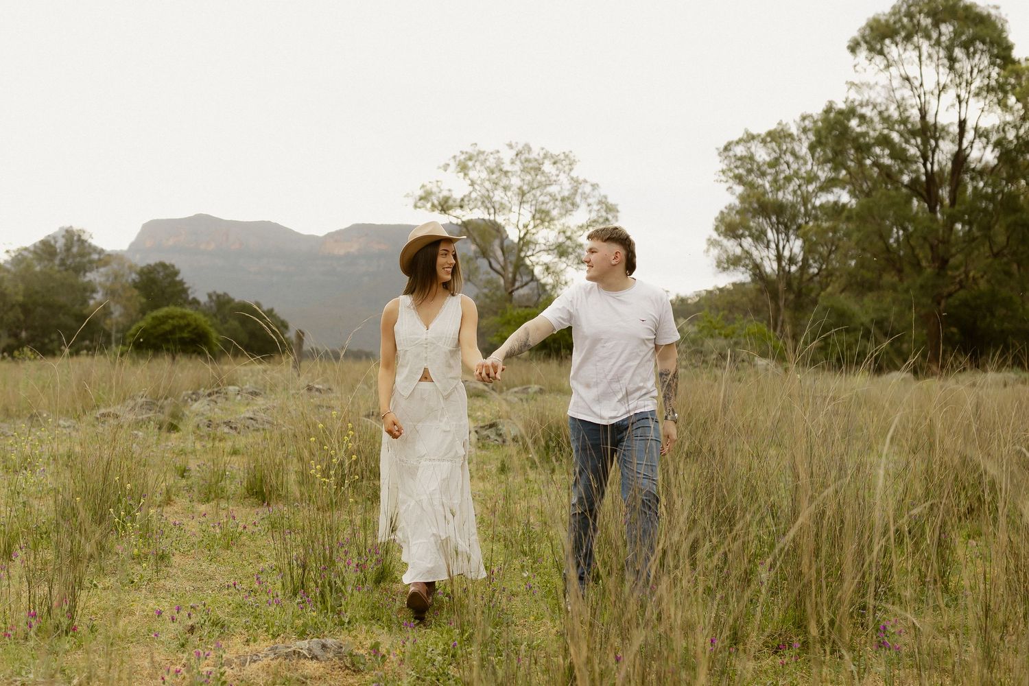 A couple walks hand in hand through a golden grass field at sunset with mountains in the background.