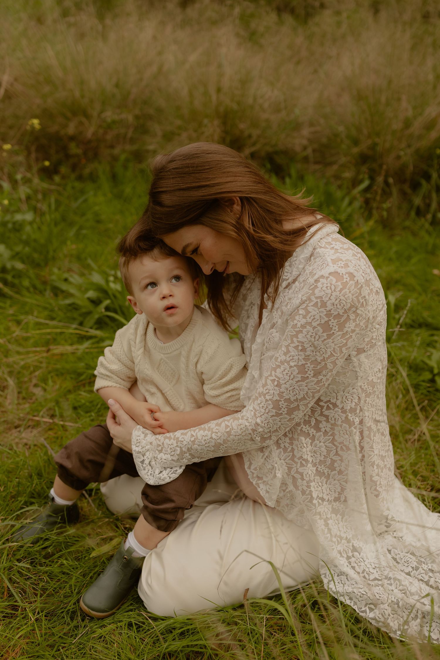 A mother in white lace holds her toddler son while sitting in a lush green meadow.