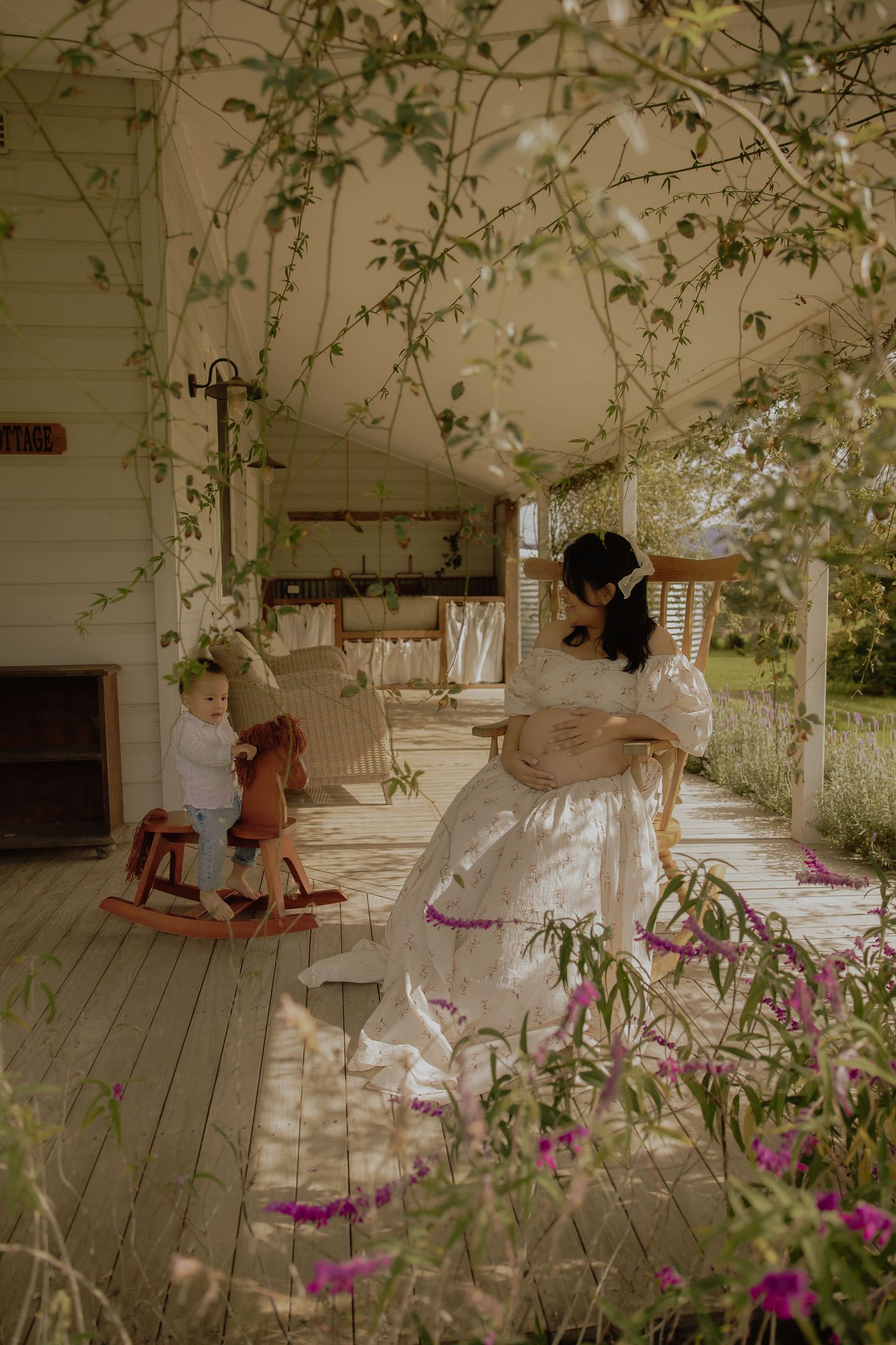 Pregnant woman in white dress sits on rustic porch while toddler plays on rocking horse nearby.