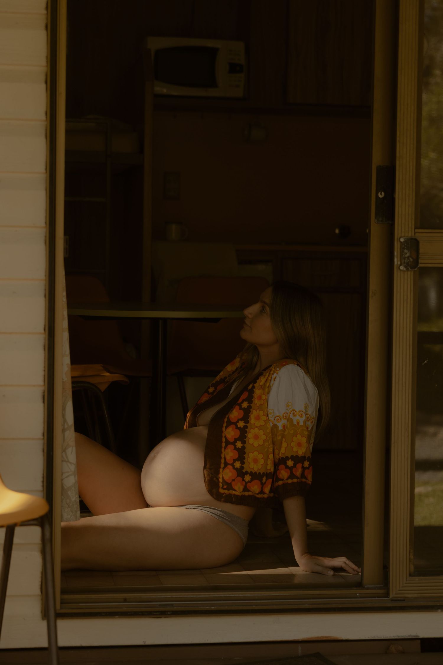 Pregnant woman in floral jacket sits on floor in doorway, bathed in warm golden light.
