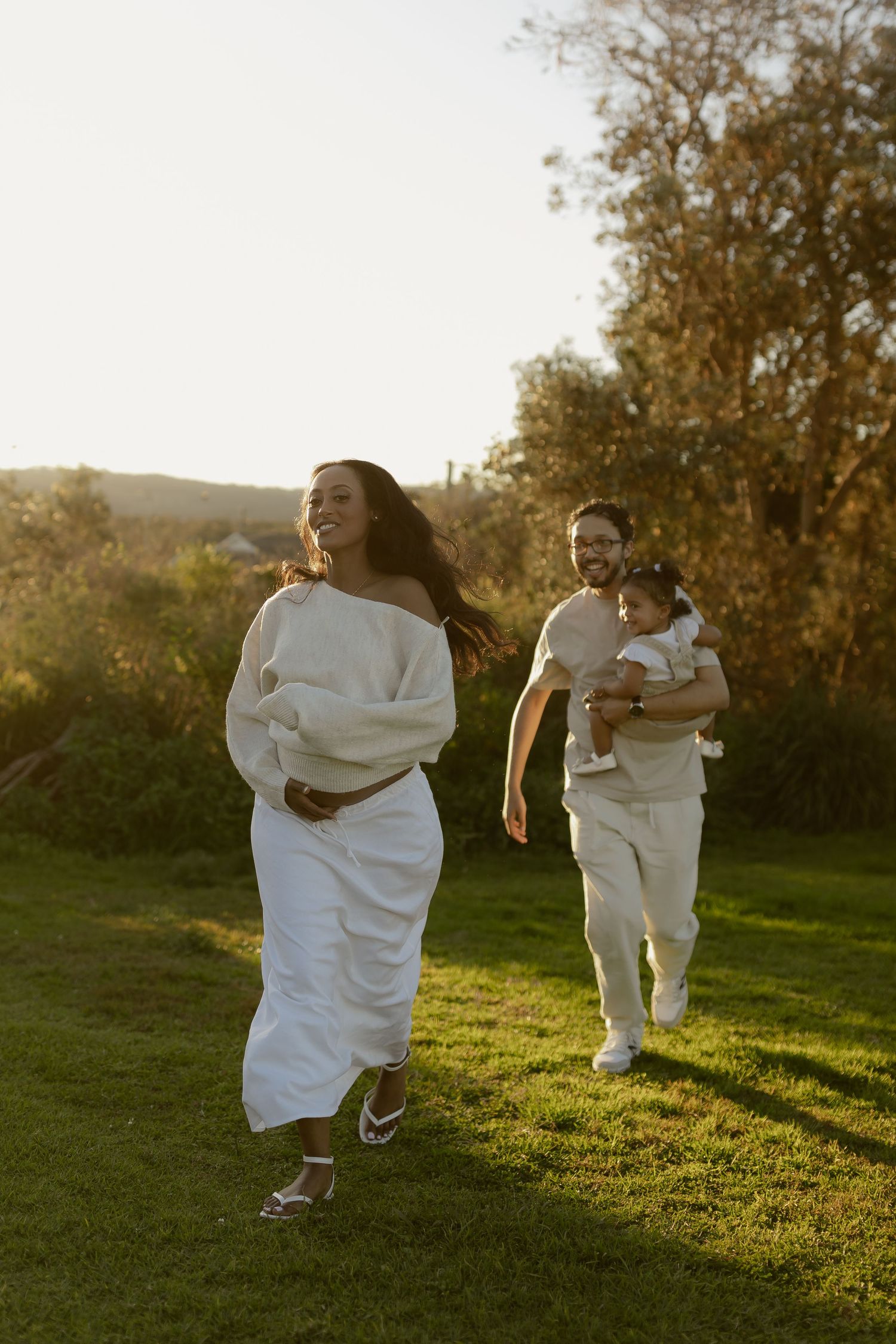 Pregnant woman in white dress runs laughing with husband carrying toddler on sunny green lawn at golden hour.