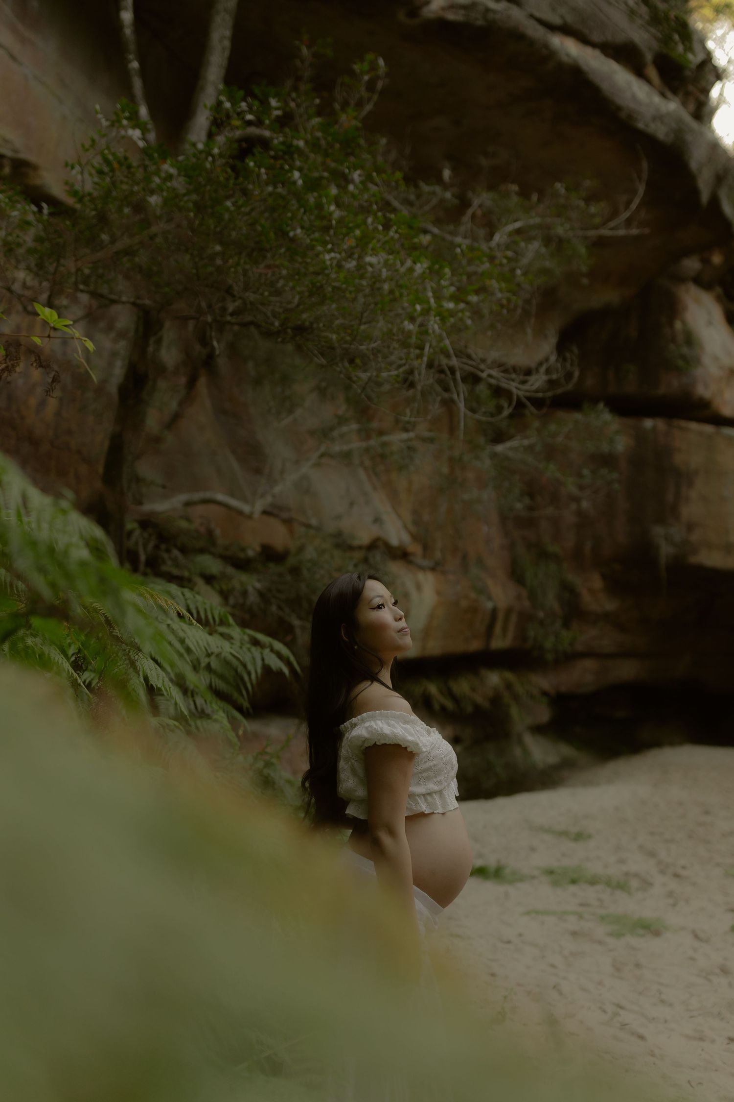 Pregnant woman in white off-shoulder top poses gracefully against sandstone rocks surrounded by ferns.