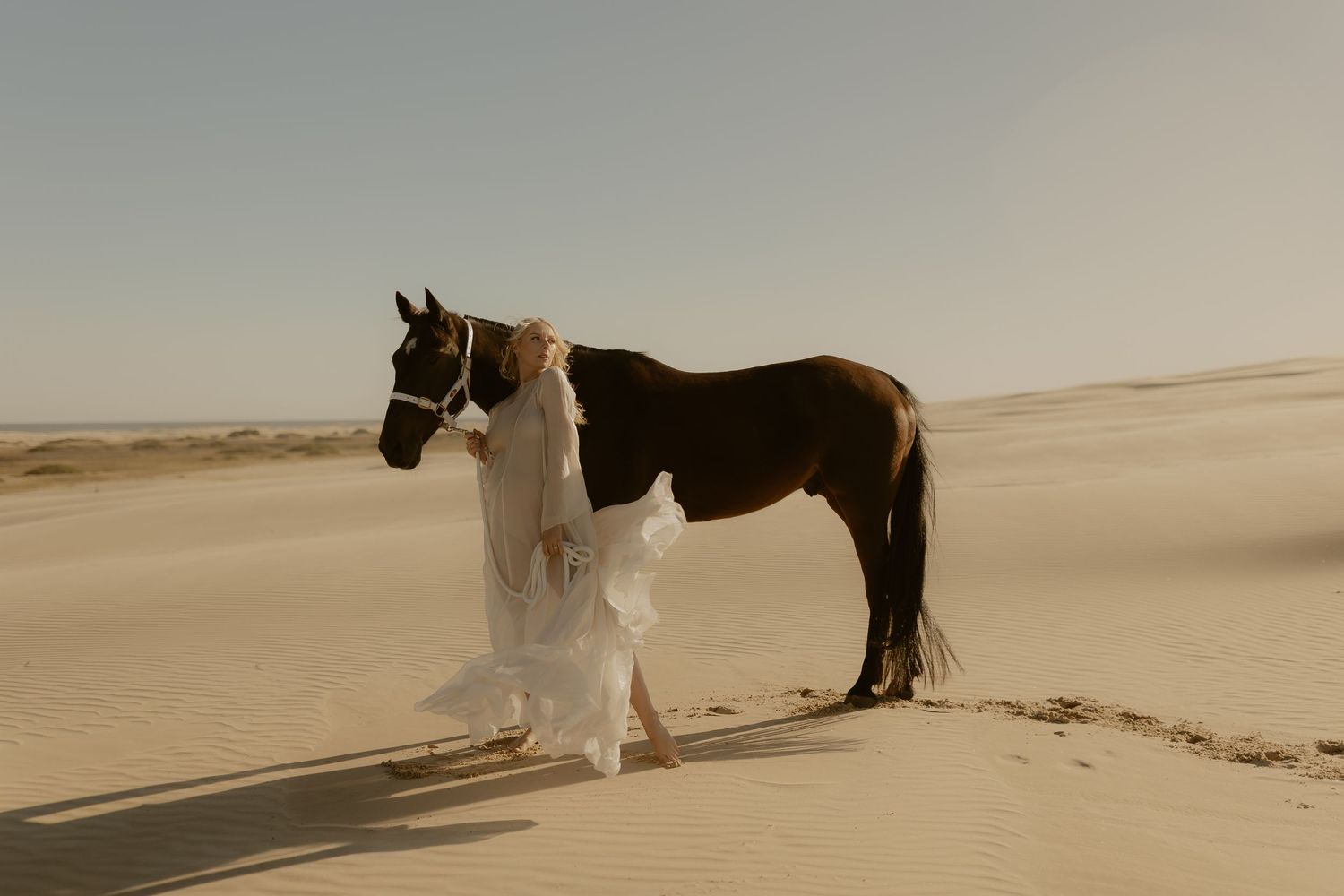 A bride in a flowing white gown stands beside a chestnut horse on golden desert sand dunes under a clear sky.
