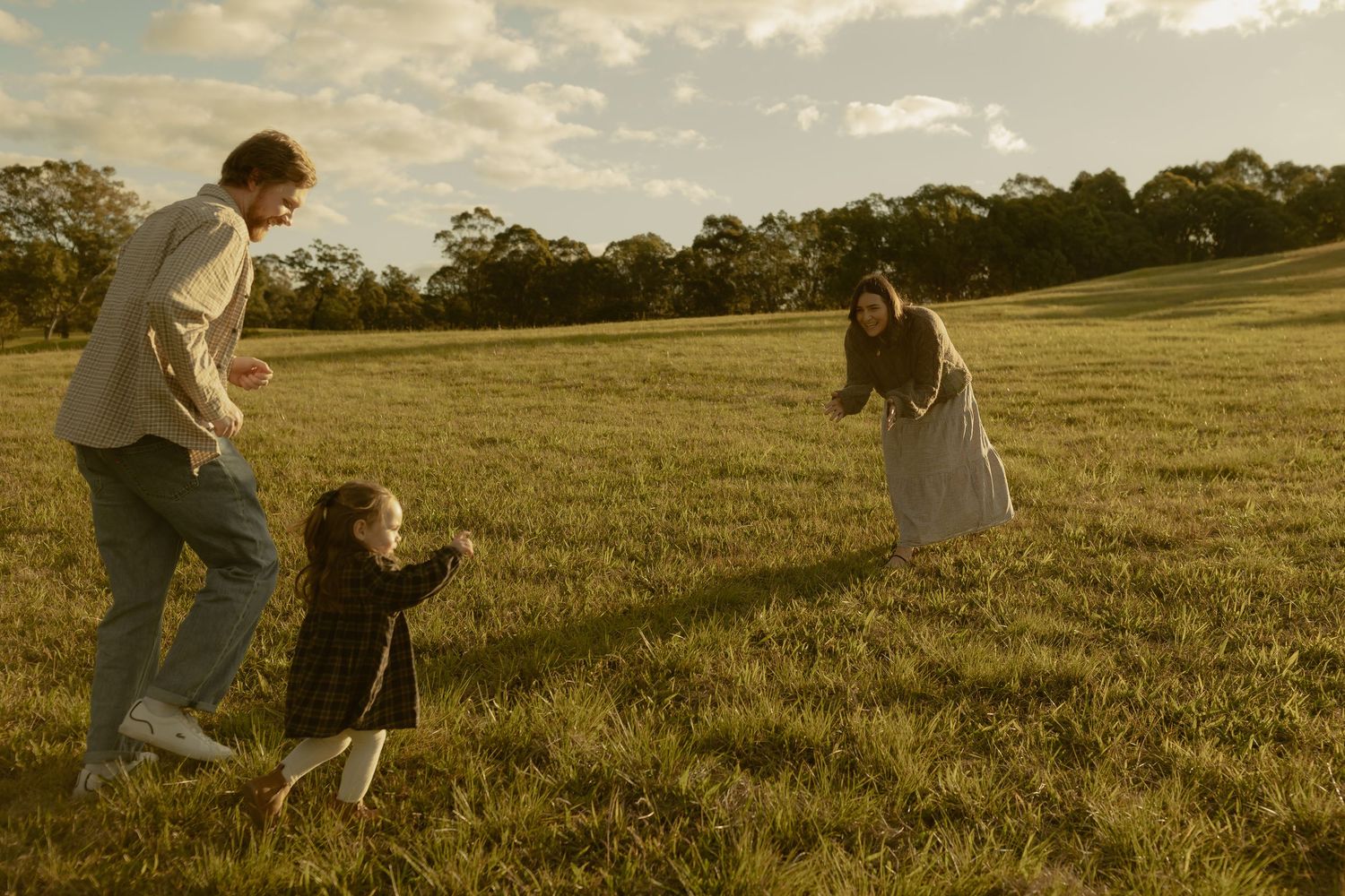Family playing in a sunlit green field at golden hour, parents encouraging toddler to walk between them.