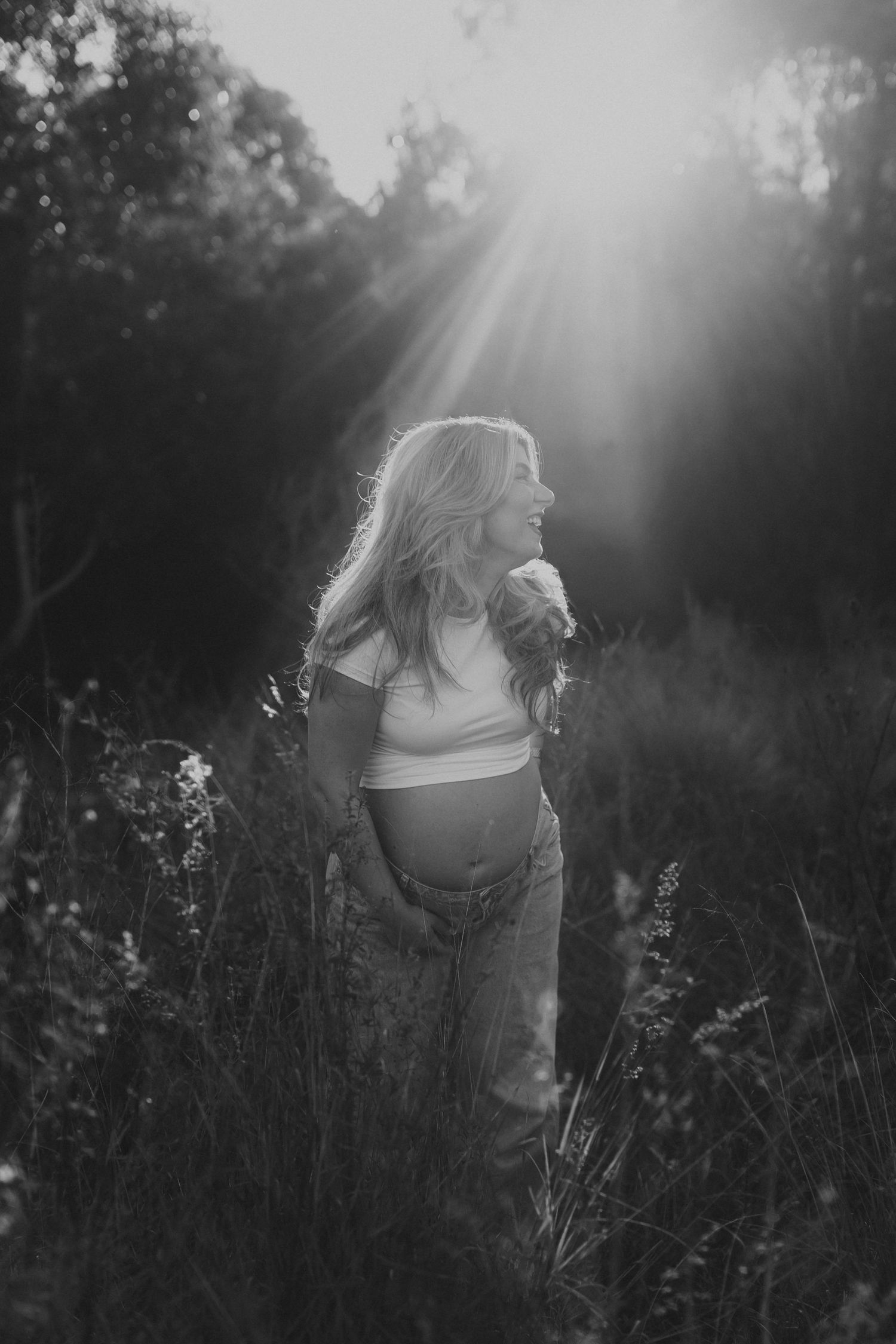 Black and white maternity photo of pregnant woman standing in tall grass with sunbeams shining through trees behind her.