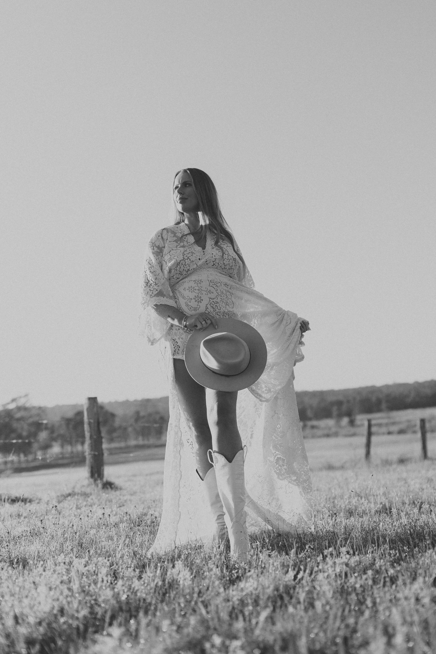 Woman in white lace dress holding hat in black and white field photo.