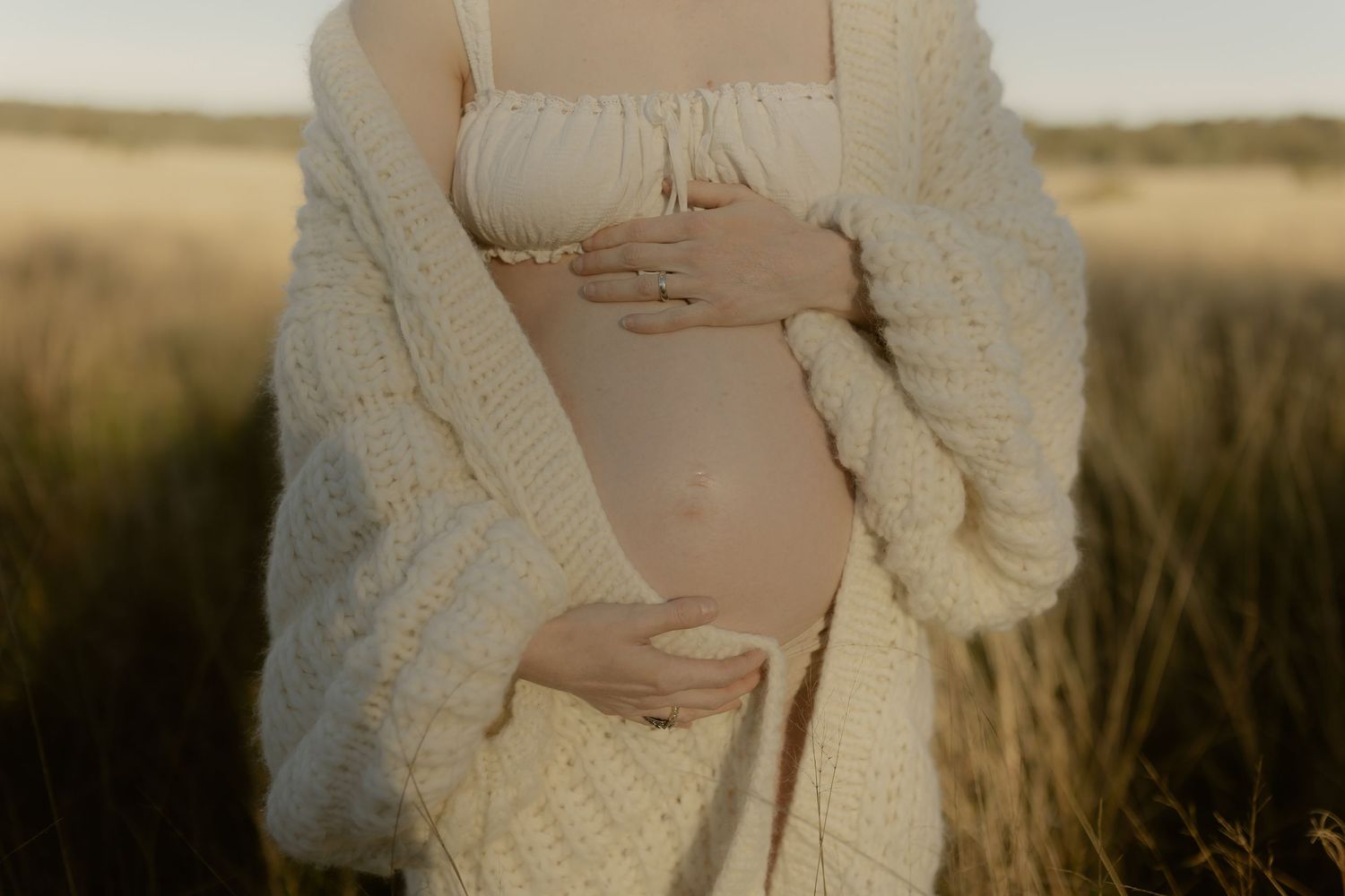 Pregnant woman in cream knit cardigan cradling bare baby bump in golden field during maternity photoshoot.