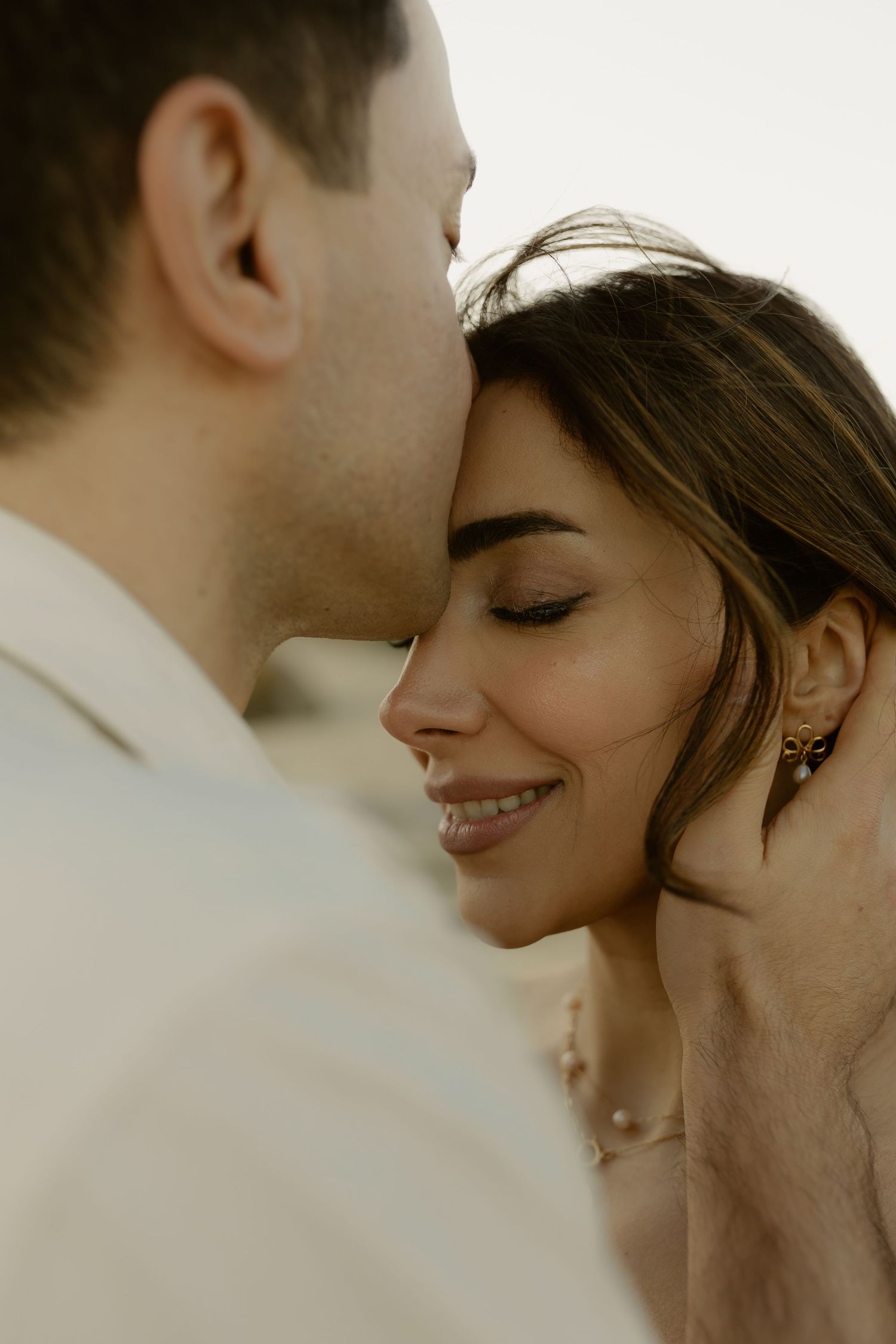 Man kissing woman's forehead as she smiles softly, wearing pearl earrings in close-up shot.