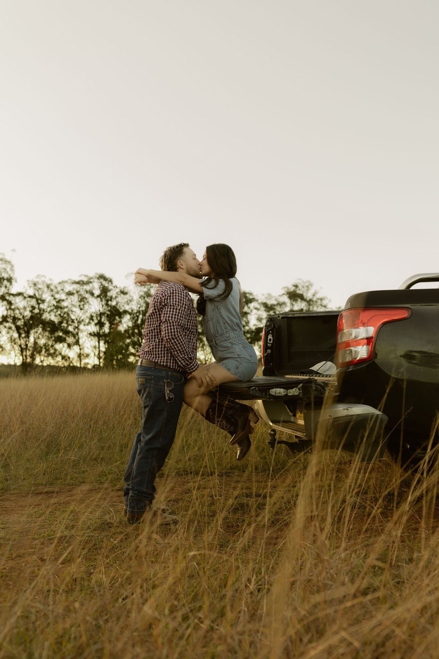 Romantic couple kissing beside a pickup truck in a rustic golden grass field at dusk.